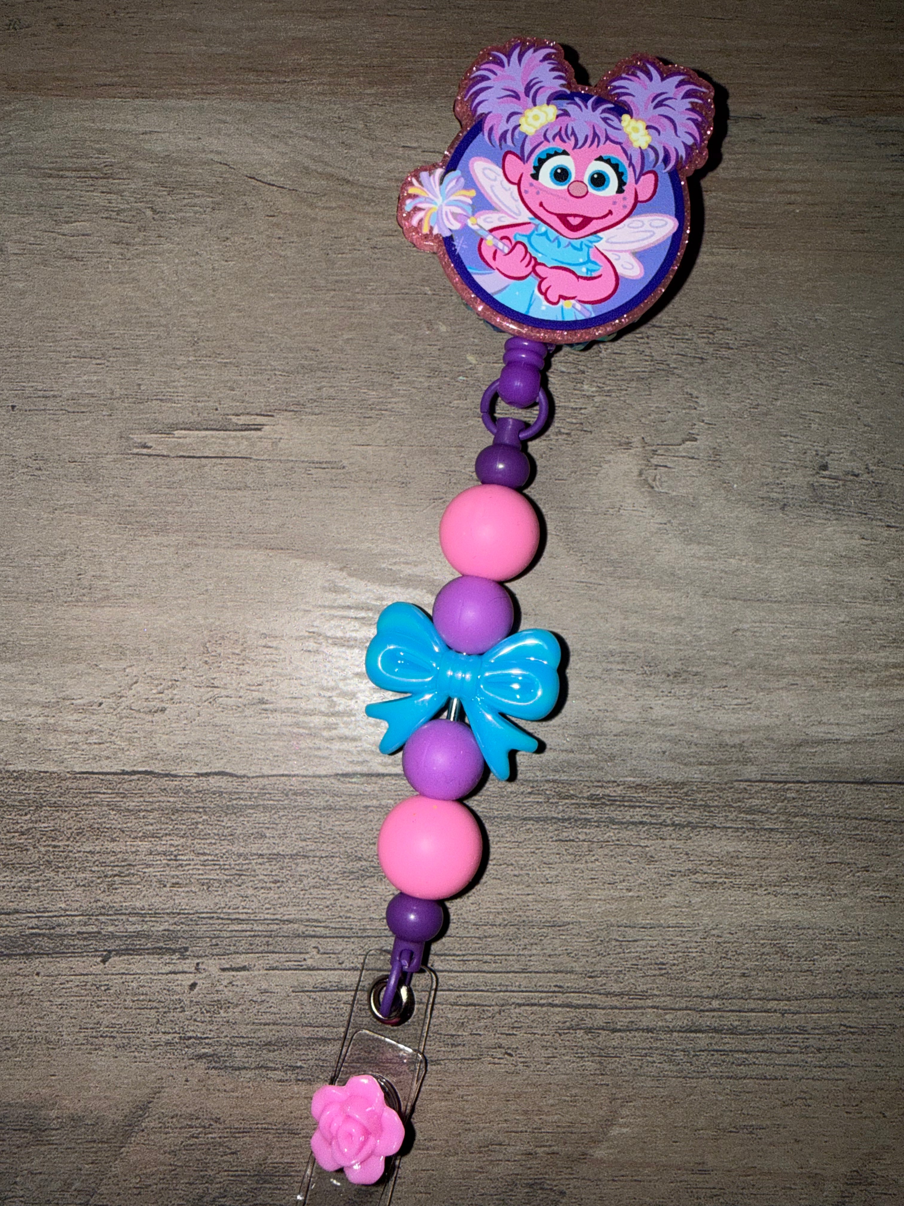 A handcrafted retractable badge reel featuring a glittery Abby Cadabby charm. The reel is decorated with a strand of purple and pink silicone beads, a bright blue bow, and a small pink rose accent at the bottom clip.