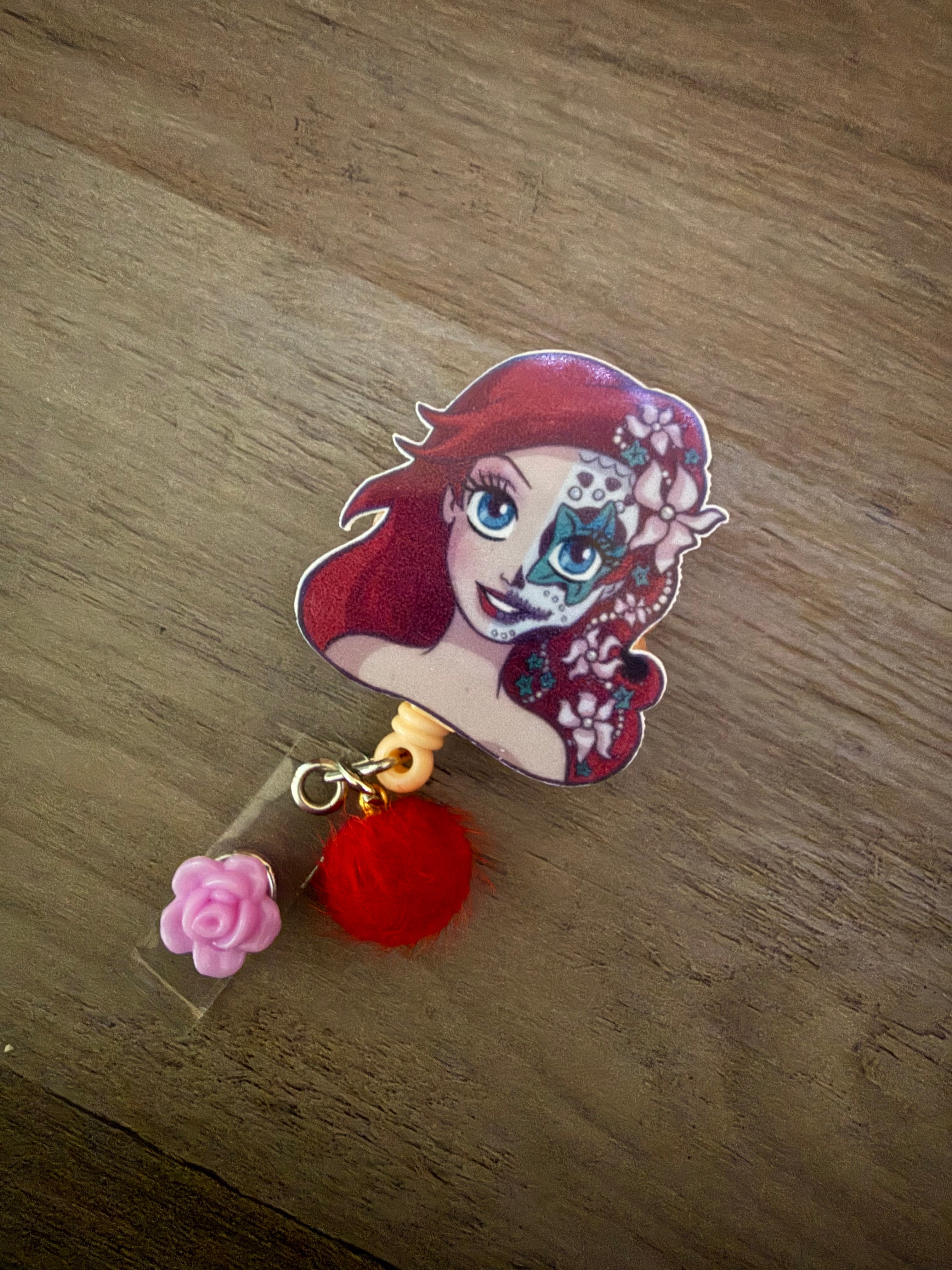 A handcrafted retractable badge reel featuring a stylized illustration of Ariel with sugar skull face paint and flowers in her hair. The reel is adorned with a bright red fuzzy pom-pom and a small lavender rose charm.