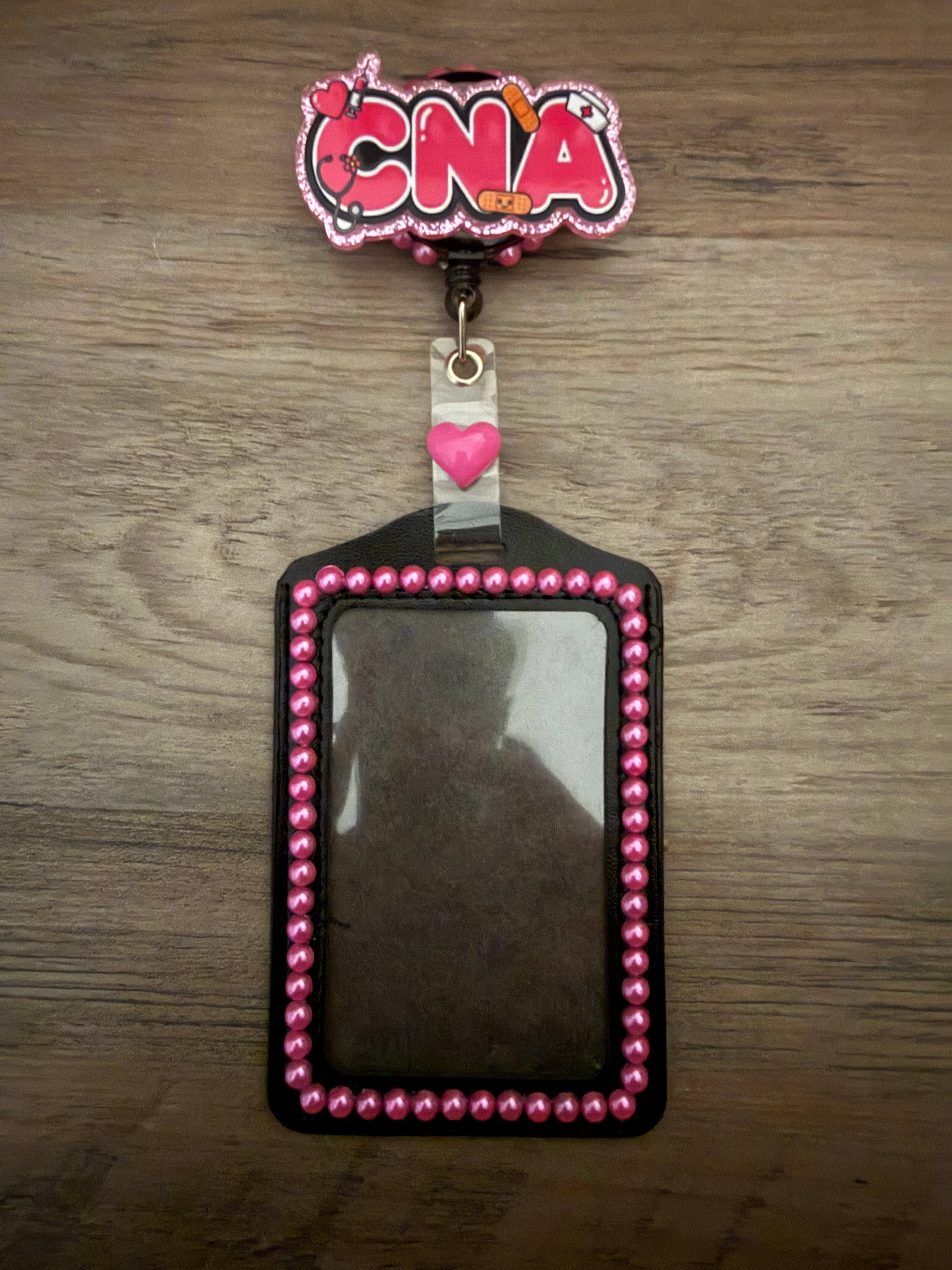A black ID badge holder with pink beaded trim, featuring a "CNA" badge reel with medical icons and a pink heart accent.
