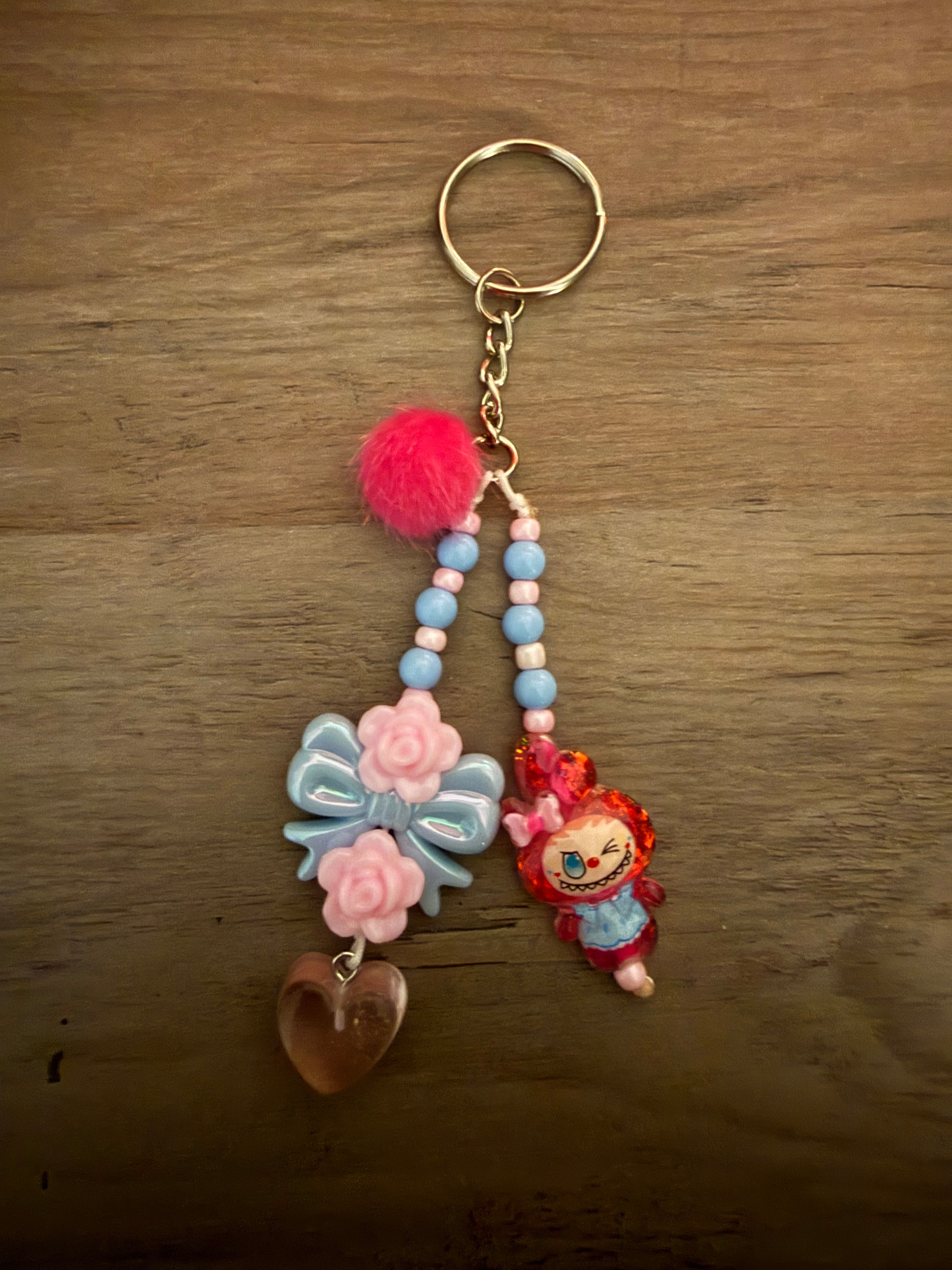 A beaded keychain featuring a hot pink pom-pom, light blue and pink beads, an iridescent blue bow with pink roses, a brown heart, and a small red clown monster charm.