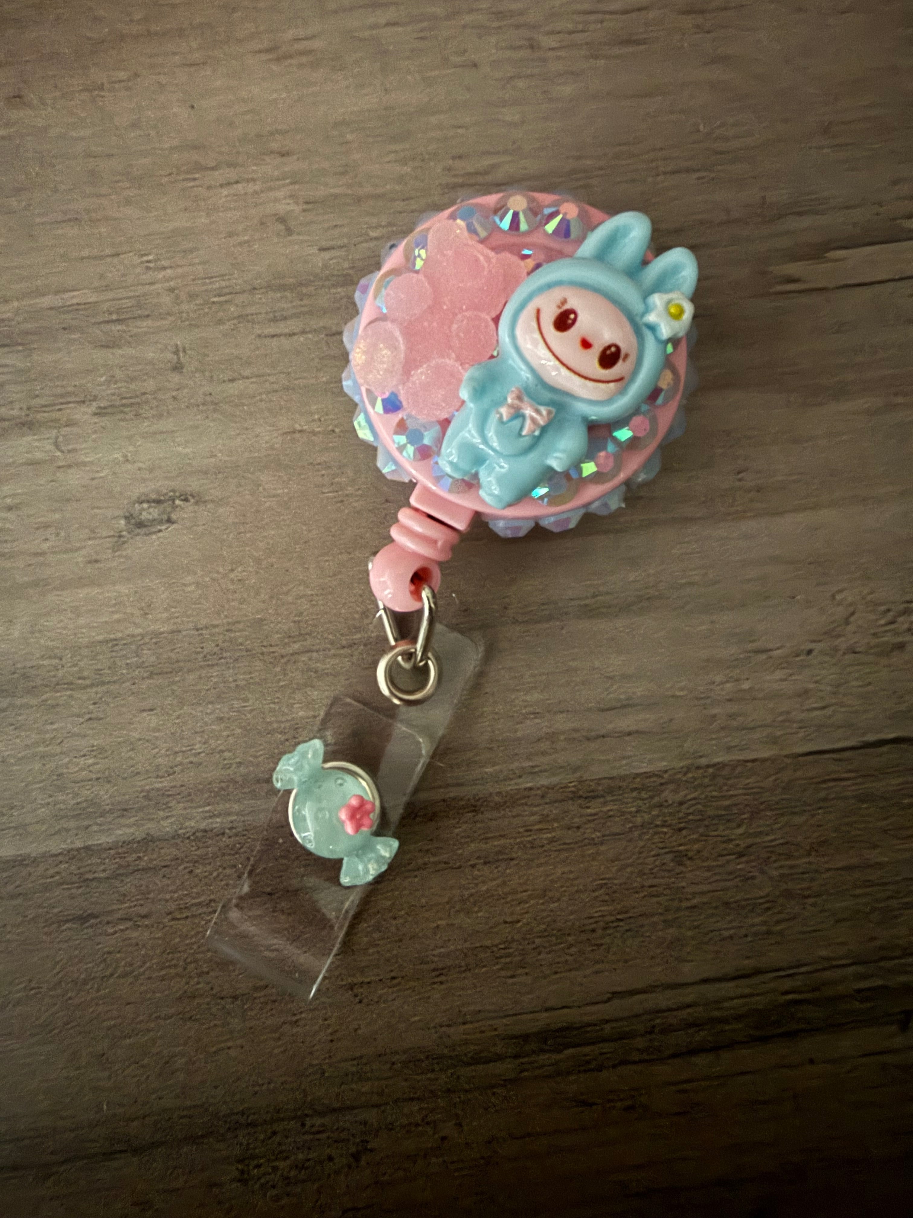 Handcrafted light blue monster in a hooded outfit on a light pink retractable badge reel, featuring iridescent rhinestones, a pink gummy bear charm, and a light blue candy charm on the strap.