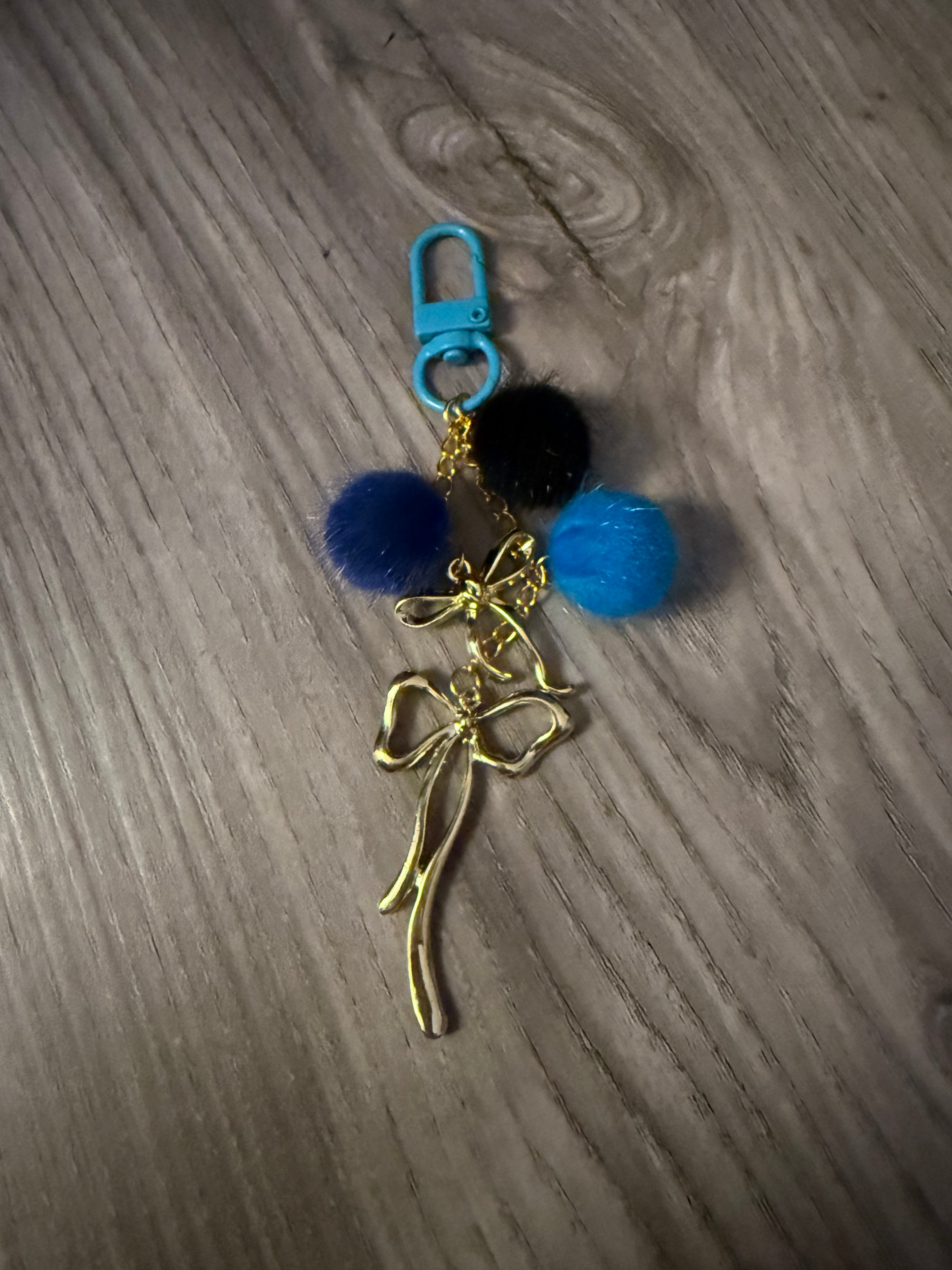 A handcrafted bag charm with a light blue lobster clasp on a wood-grain surface. The charm features a gold-tone chain adorned with three small pom-poms in dark blue, black, and bright blue, finished with two elegant gold-tone metal bows.