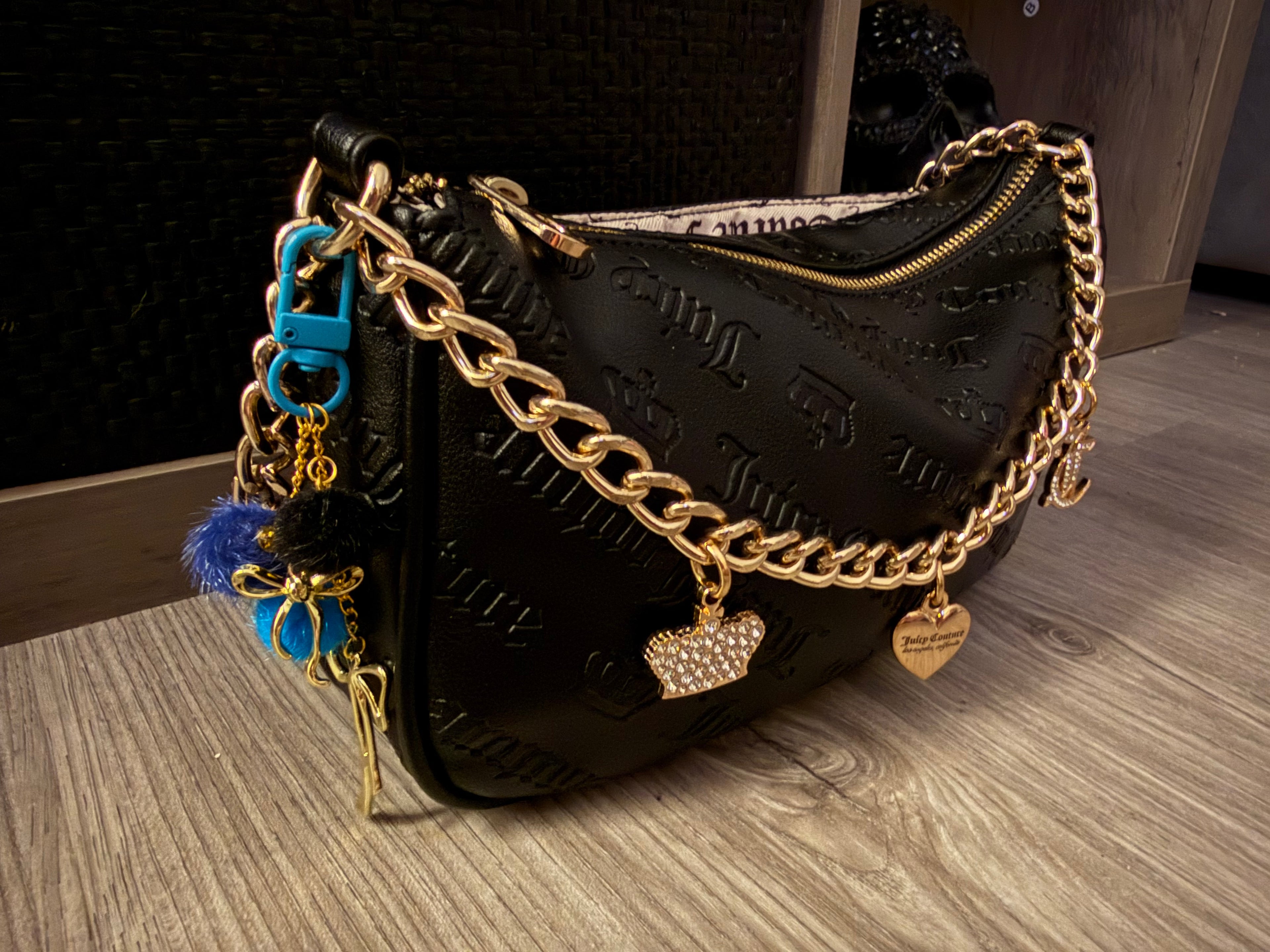 A black embossed Juicy Couture handbag featuring a gold-tone chain strap with dangling charms, including a crystal-encrusted crown and a gold heart. A handcrafted bag charm with blue and black pom-poms and a blue lobster clasp is attached to the side of the bag.