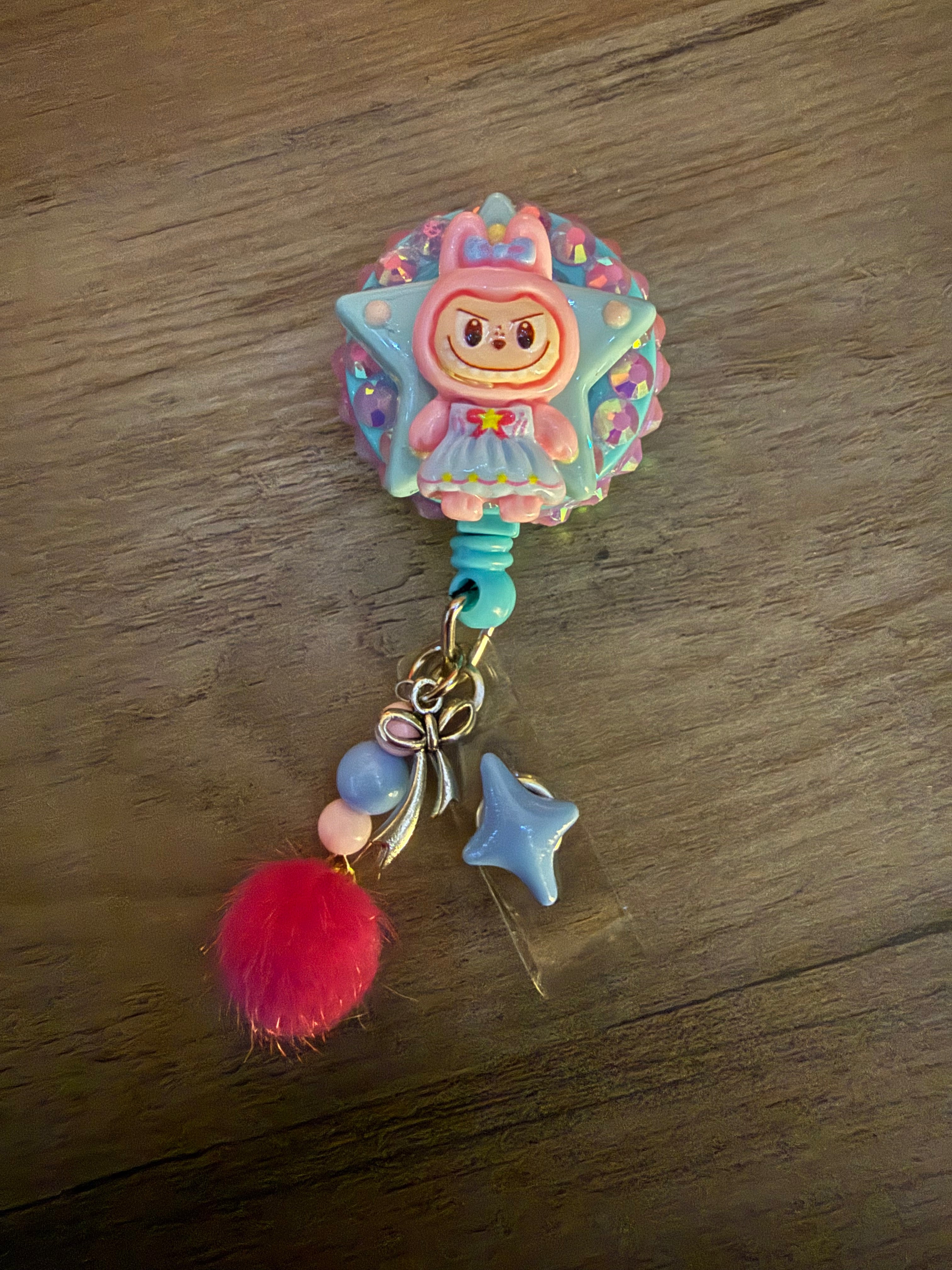A handcrafted, decoden-style badge reel featuring a pink monster in a blue dress set against a glittering, rhinestone-encrusted star. The reel is adorned with hanging charms including a pink pom-pom, pastel beads, a silver bow, and a blue star.