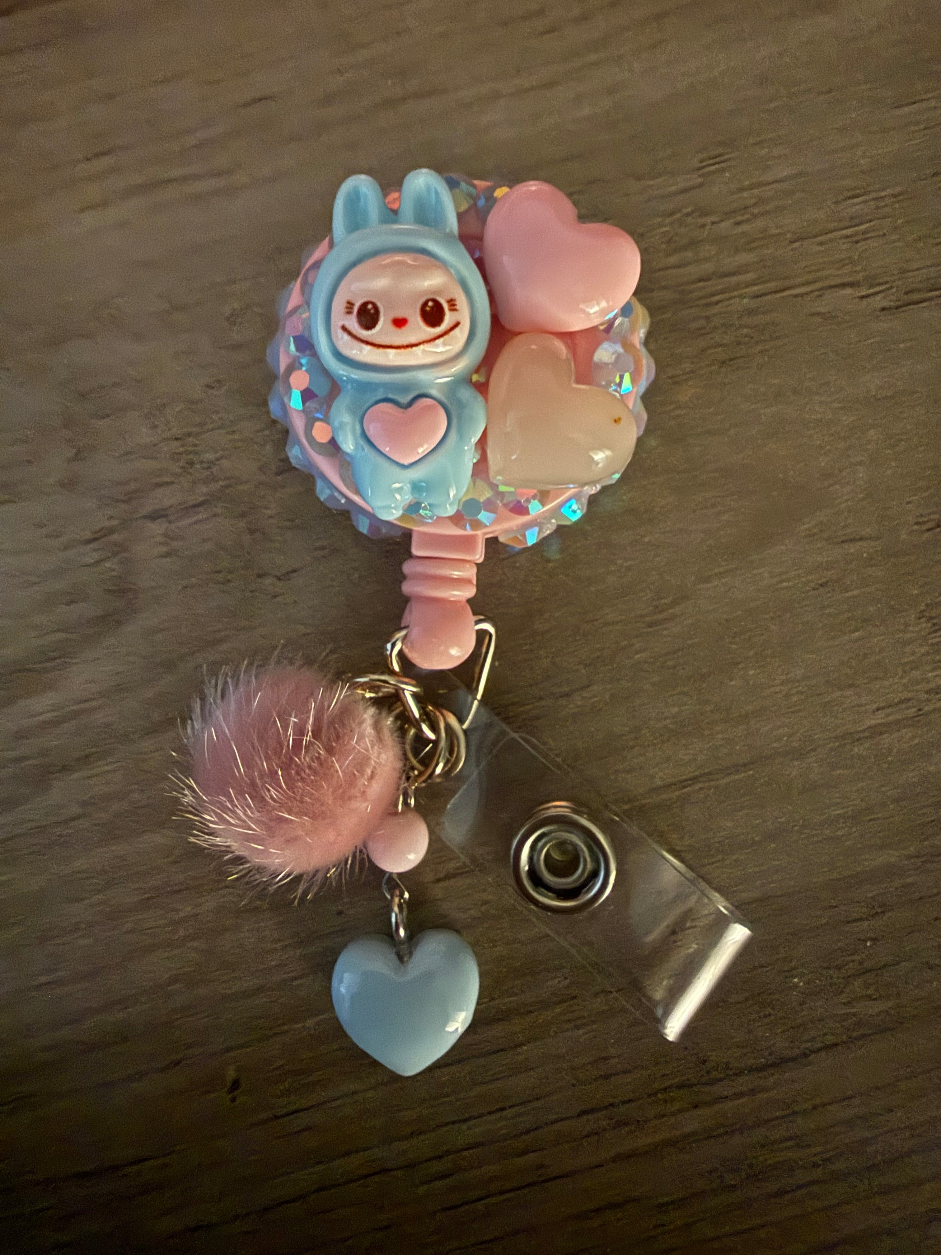 A light blue and pink handcrafted decoden badge reel featuring a small blue monster, pink resin hearts, and iridescent rhinestones, accented with a pink pom-pom and a light blue heart charm.