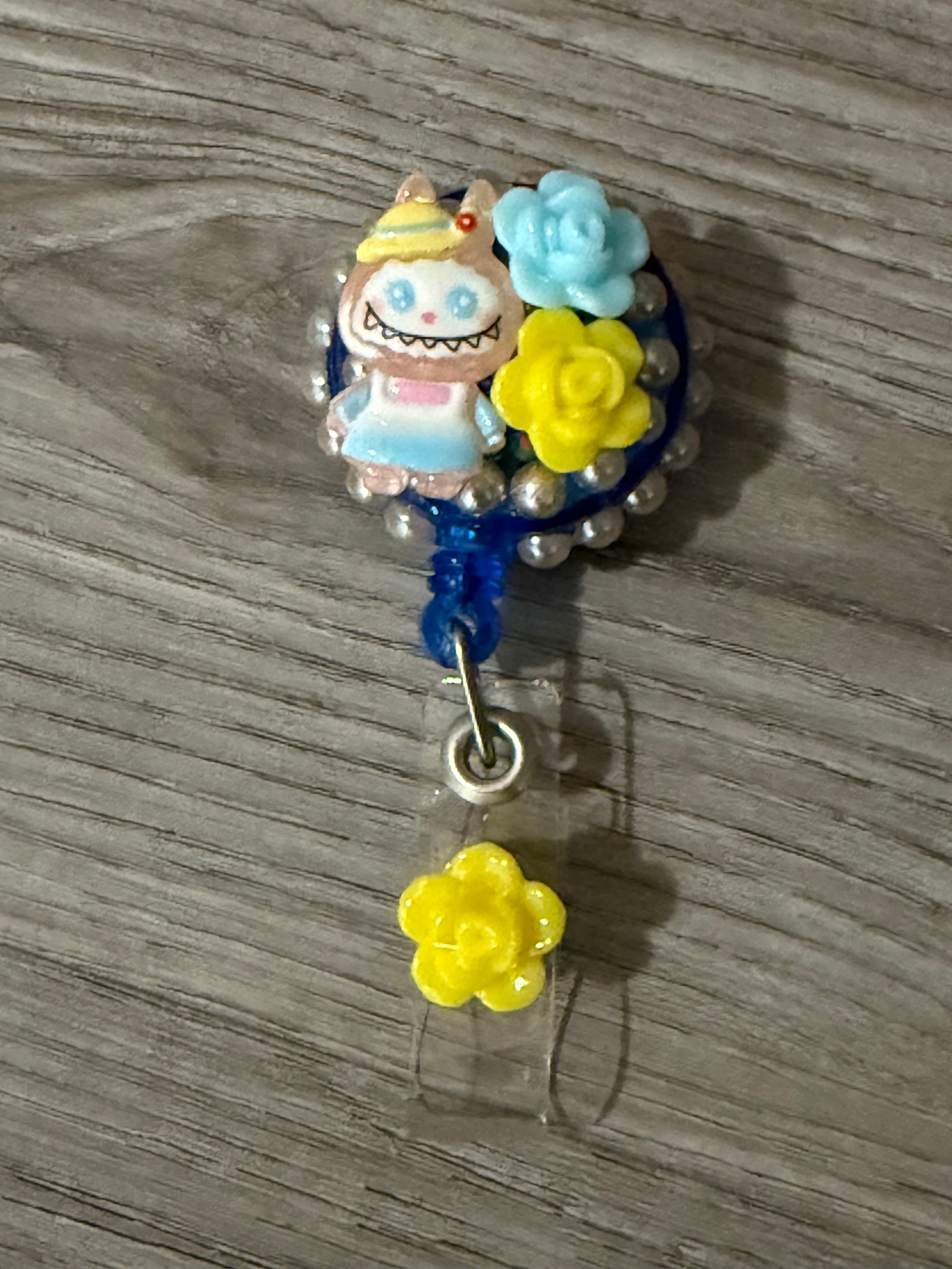 Cute badge reel featuring a whimsical monster with bunny ears and a playful grin, adorned with blue and yellow flowers, surrounded by pearl beads.