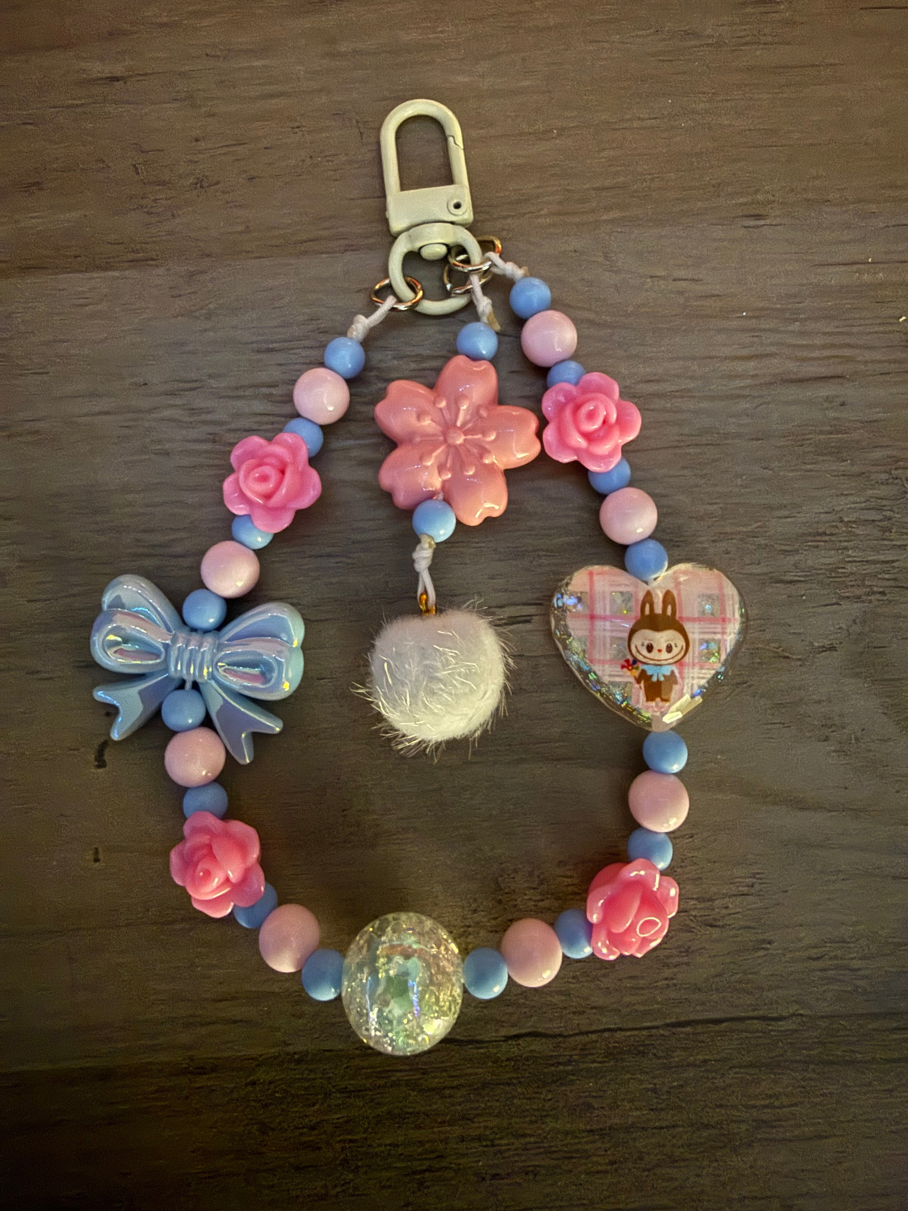 A handcrafted pink and blue beaded bag charm featuring rose accents, a blue bow, a white pom-pom, and a heart-shaped monster pendant.
