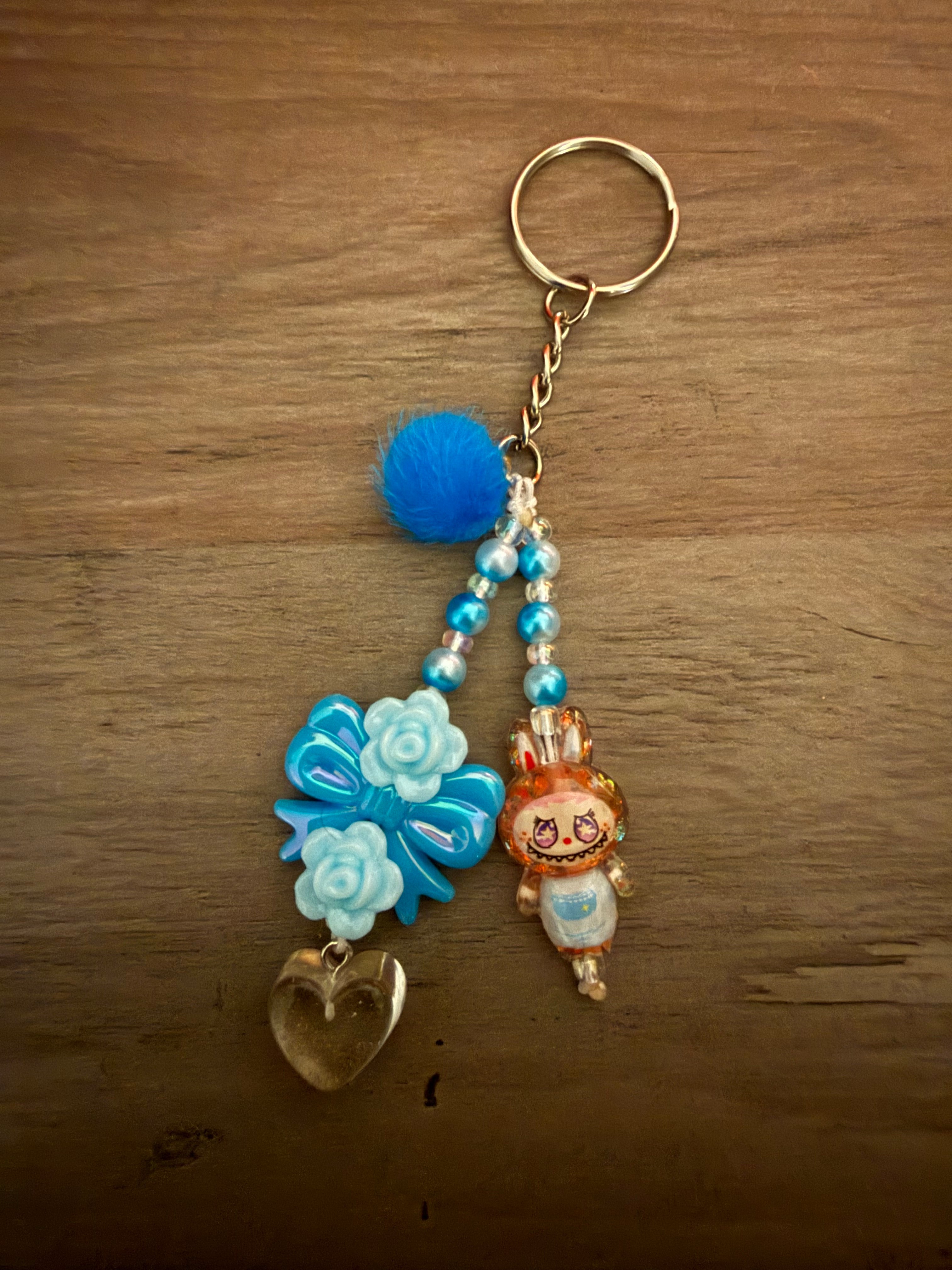 A handcrafted blue keychain featuring two beaded strands. The design includes a fluffy blue pom-pom, a cartoon monster character in a blue outfit, a bright blue bow accented with light blue roses, and a clear heart-shaped charm.