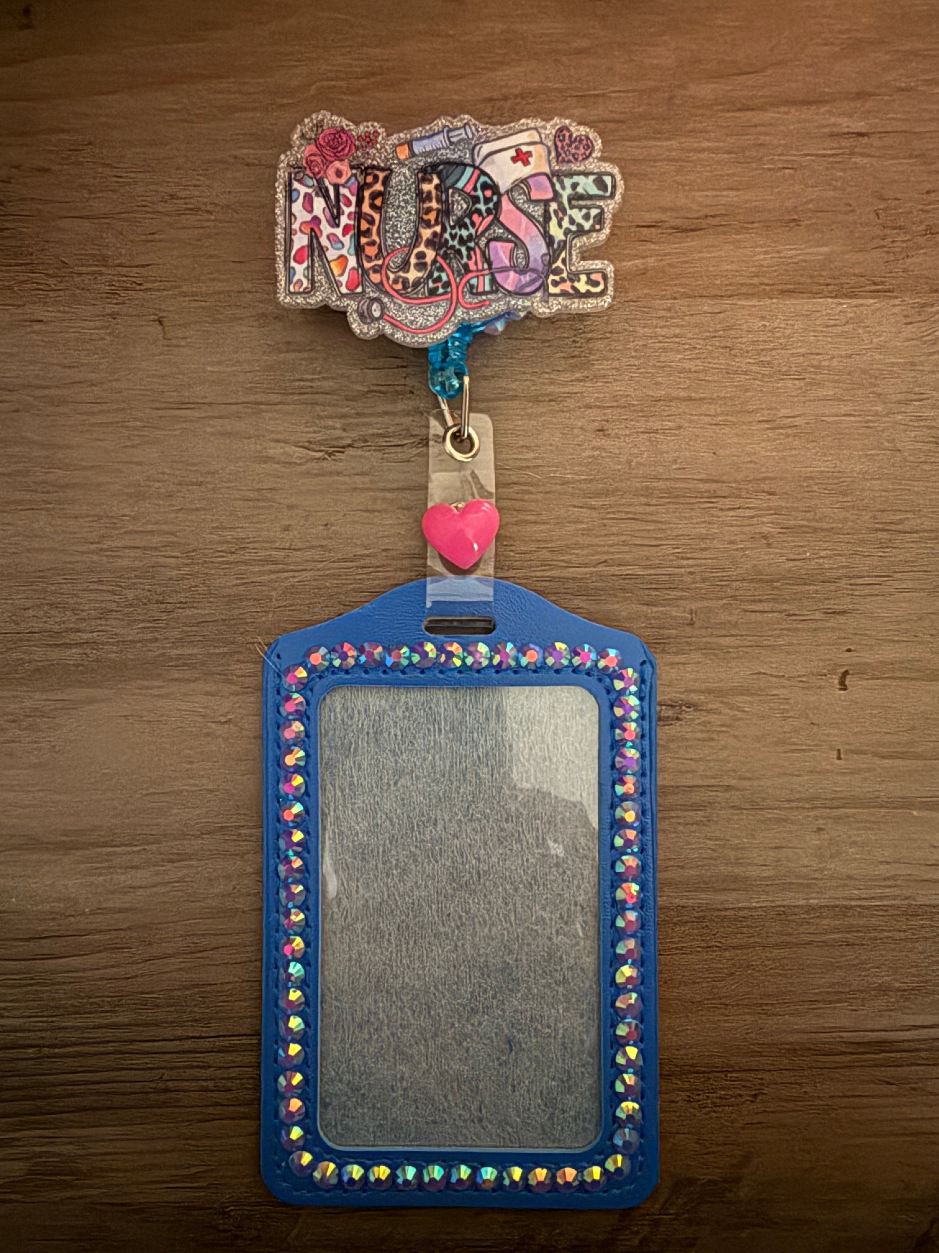 A blue, rhinestone-lined ID holder attached to a glittery badge reel featuring the word "NURSE" in a colorful leopard and floral print. A small pink heart charm accents the clip.