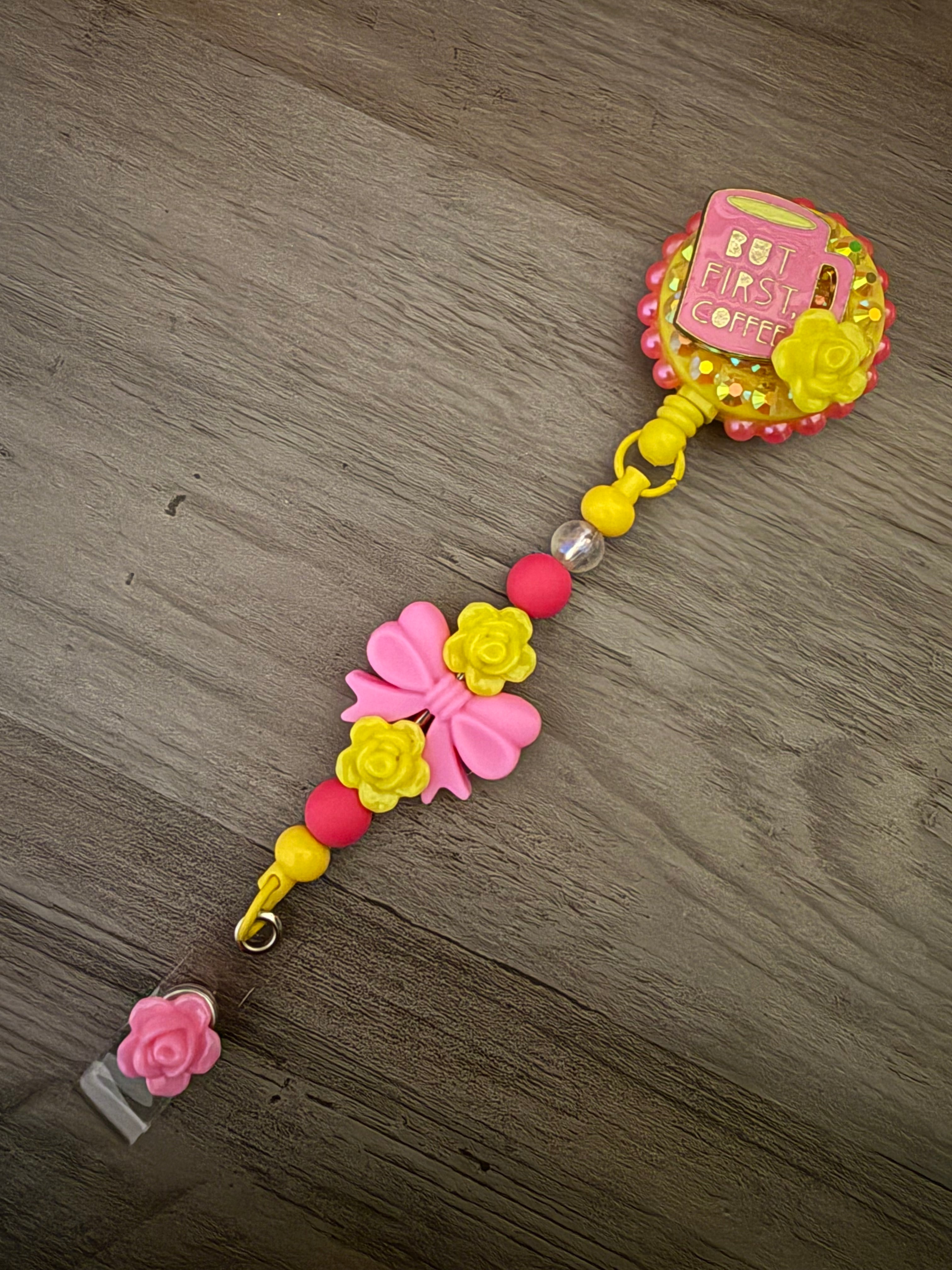 Handmade yellow badge reel featuring a pink coffee mug charm with the text "But First, Coffee" on a glittery base, accented by a decorative lanyard with pink bows, yellow rose beads, and a pink flower dangle.