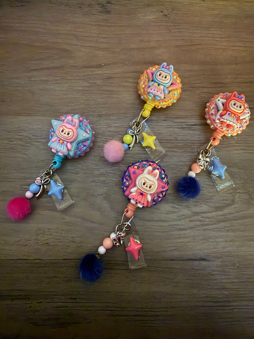 Four handmade, decoden-style retractable badge reels featuring colorful monster on star-shaped bases. The reels are decorated with glitter, rhinestones, and coordinating charms including pom-poms, beads, and bows in pastel pink, blue, and yellow.
