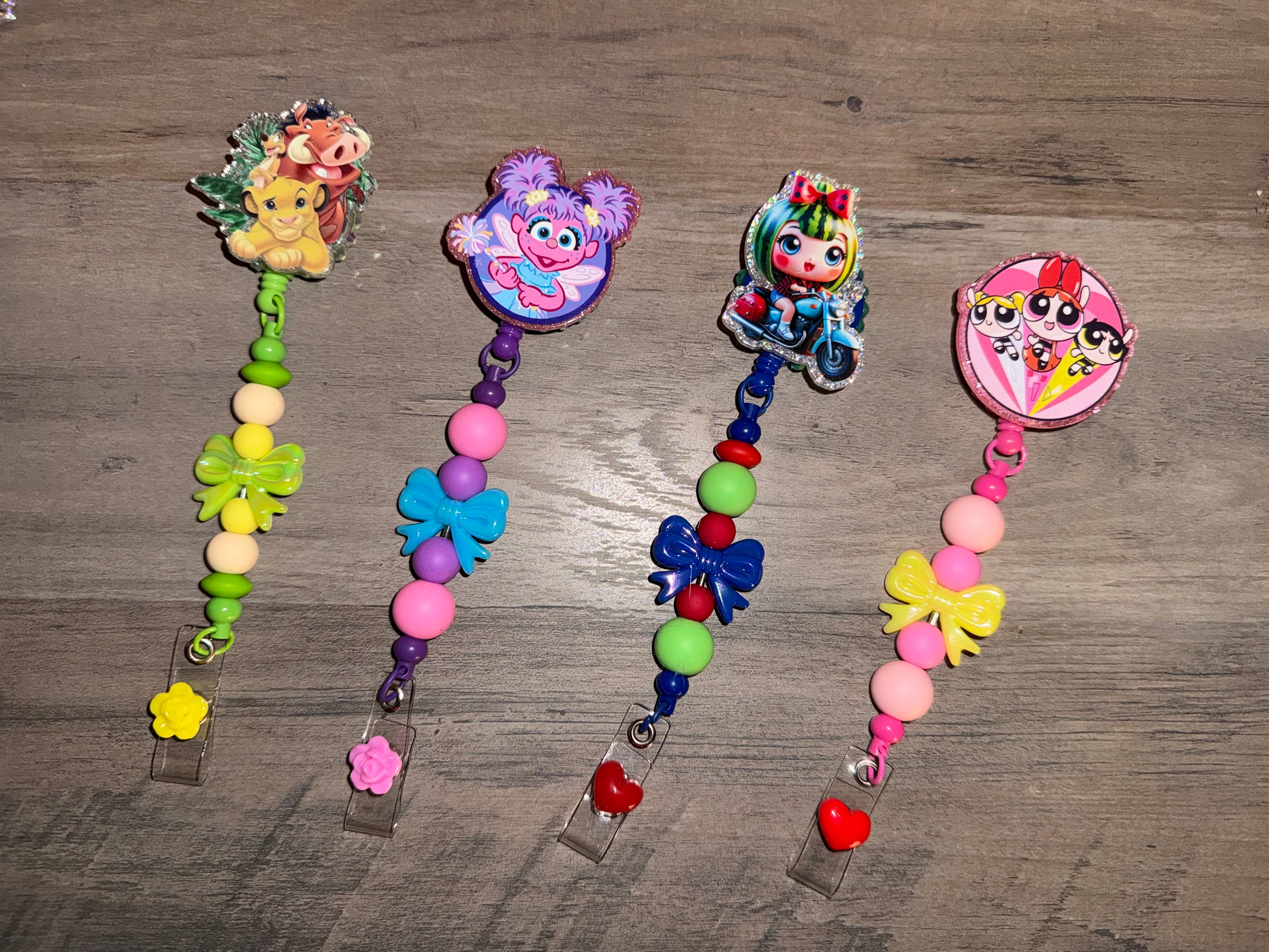 Four handcrafted, beaded retractable badge reels featuring colorful character charms including Simba, Timon, and Pumbaa; Abby Cadabby; a doll on a motorcycle; and The Powerpuff Girls. Each reel is decorated with coordinating silicone beads, colorful bows, and small floral or heart-shaped accents at the base.