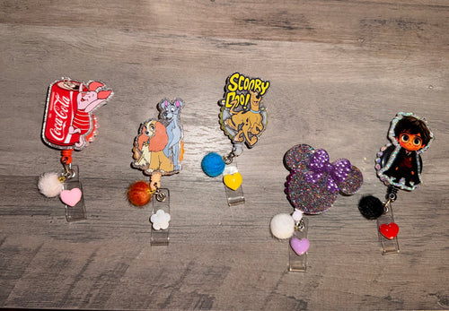 A collection of five character-themed glitter badge reels arranged in a row. From left to right: Piglet with a Coca-Cola can, Lady and the Tramp, Scooby-Doo, Minnie Mouse, and Harry Potter. Each reel is decorated with a matching color-coordinated pom-pom and a small heart charm hanging from the clip.