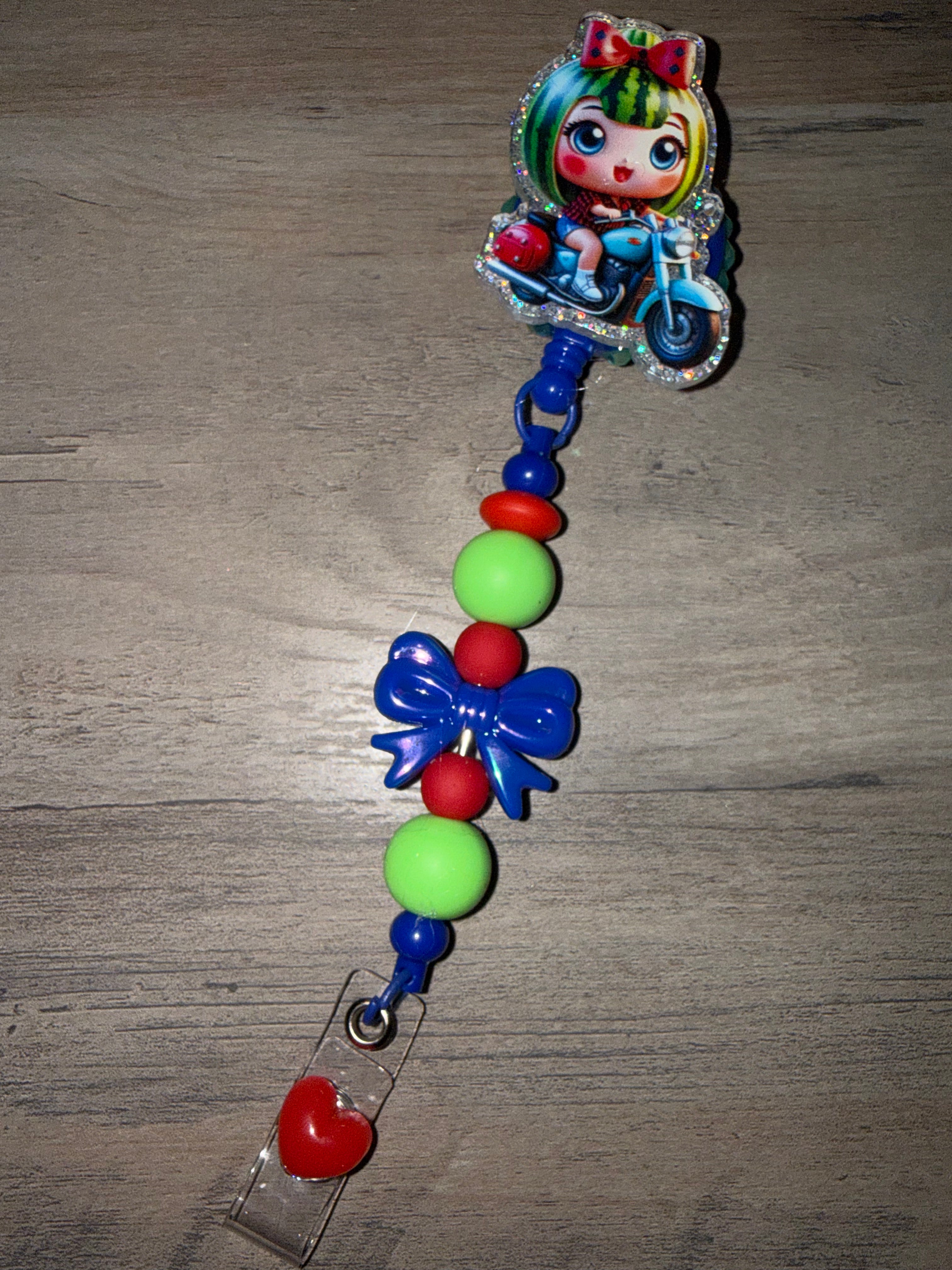 A handcrafted retractable badge reel featuring a glittery charm of a doll with watermelon-styled hair riding a motorcycle. The reel is embellished with lime green, red, and blue beads, a dark blue bow, and a red heart accent at the bottom clip.