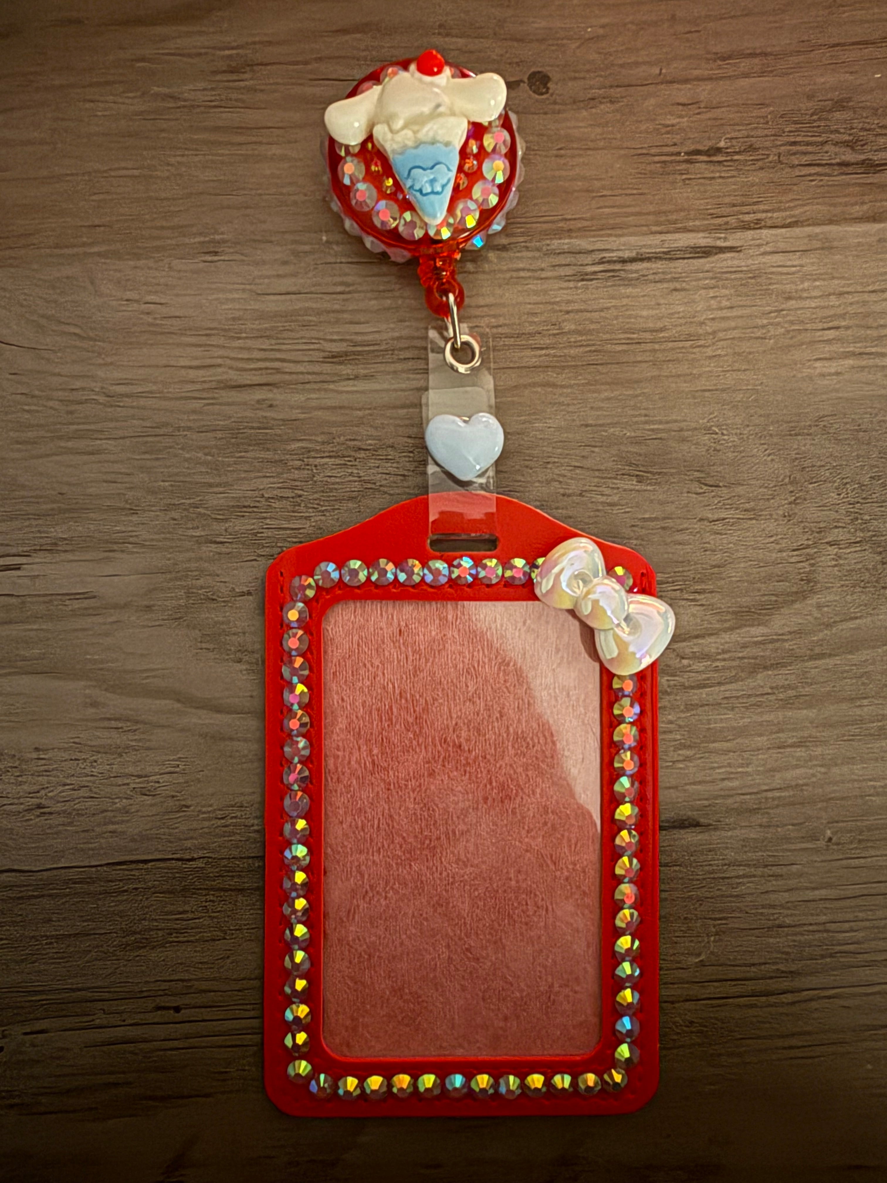 A red, rhinestone-embellished ID card holder with a matching retractable badge reel. The reel features a Cinnamoroll ice cream cone charm set against a sparkling red background, and the holder is bordered with iridescent gems and a small white pearlescent bow.