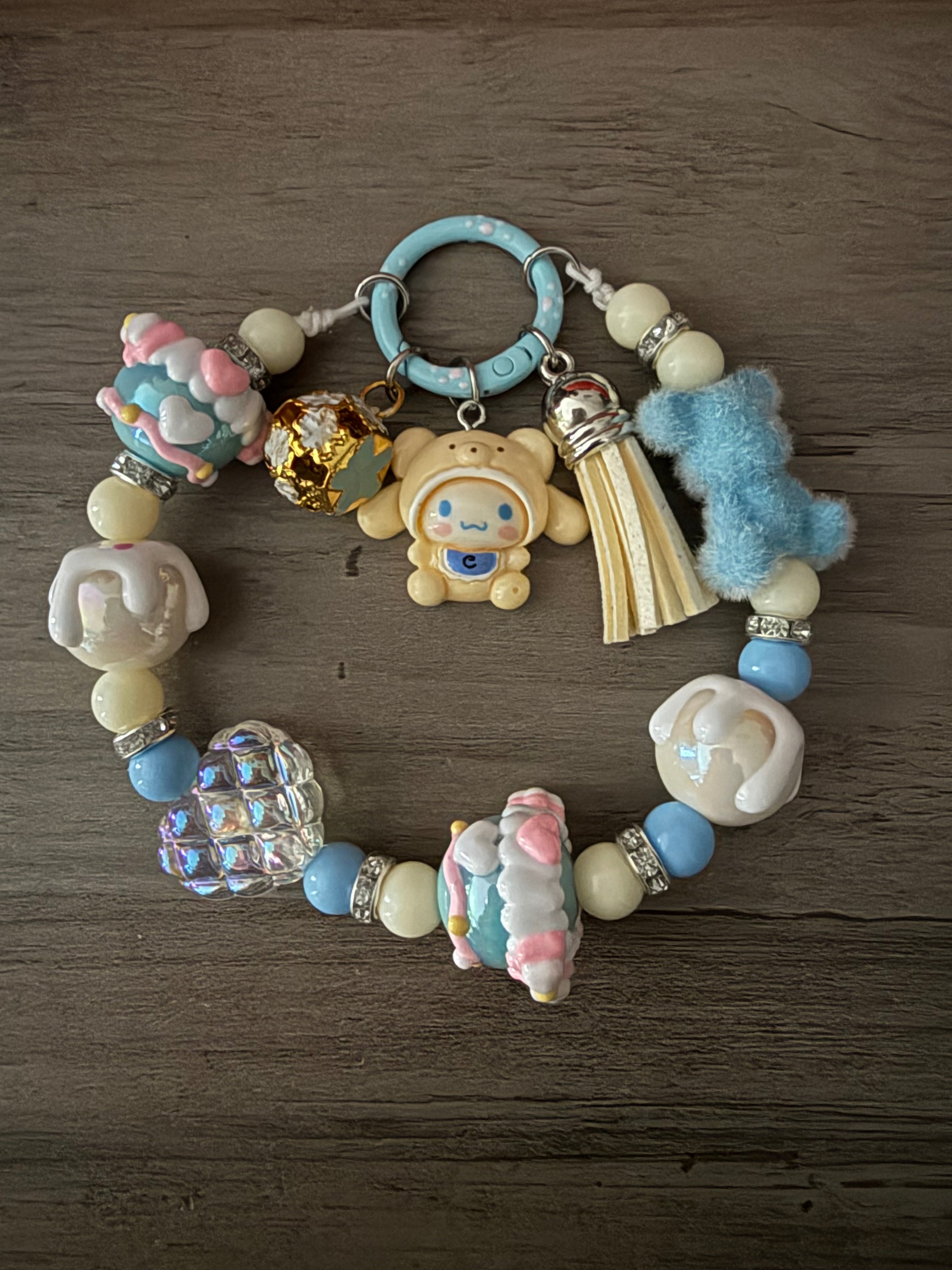 A pastel blue and cream beaded charm featuring Cinnamoroll figures, a fuzzy blue bear, and a white tassel. The beads include cream and blue rounds, silver rhinestone spacers, and an iridescent quilted heart, all connected to a light blue circular clasp.