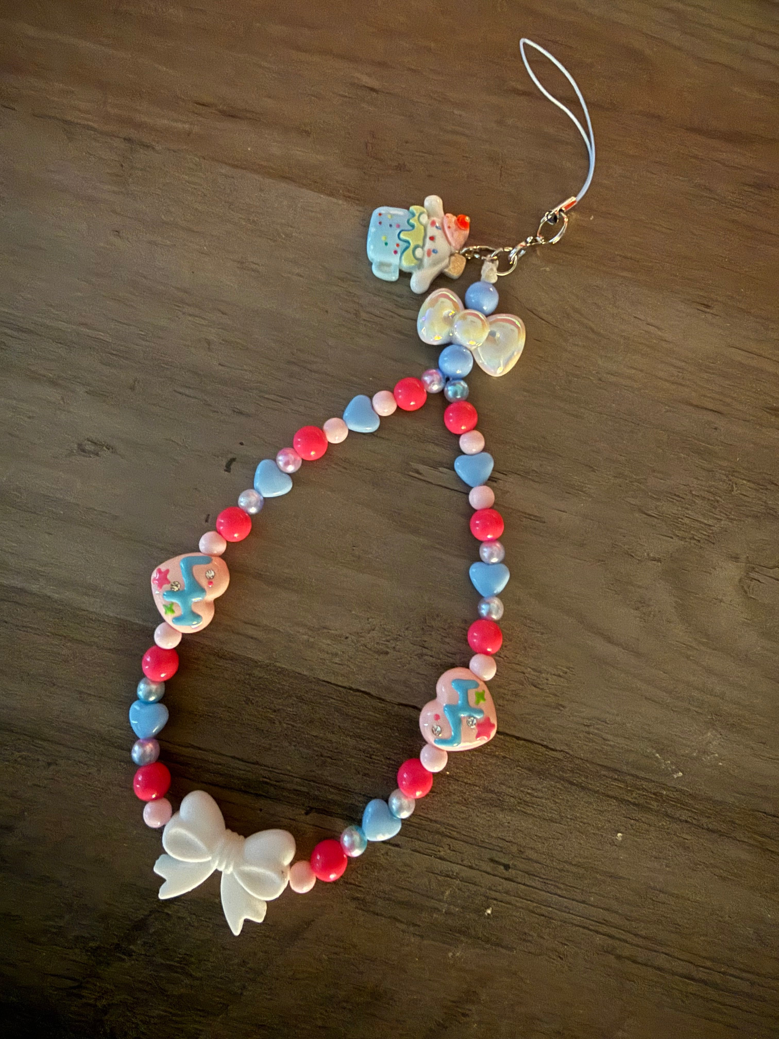 A colorful Cinnamoroll inspired, beaded phone wristlet strap featuring a mix of pink, blue, and white beads, heart-shaped charms, and white decorative bows, laid out on a dark wooden surface.