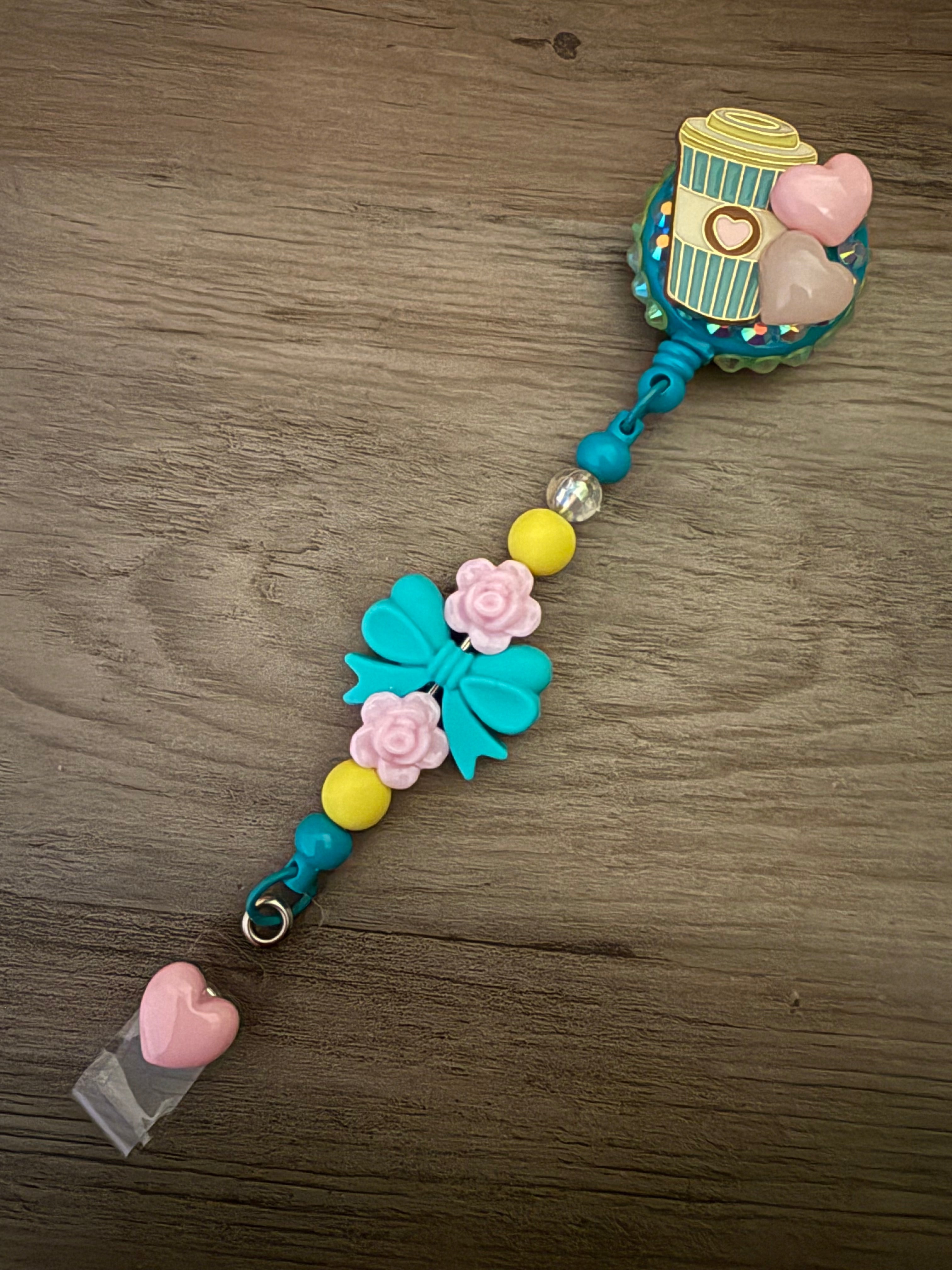 Handmade teal badge reel featuring a coffee cup charm and pink heart accents on a glittery base, with a decorative beaded lanyard including a teal bow, pink roses, and a pink heart dangle.