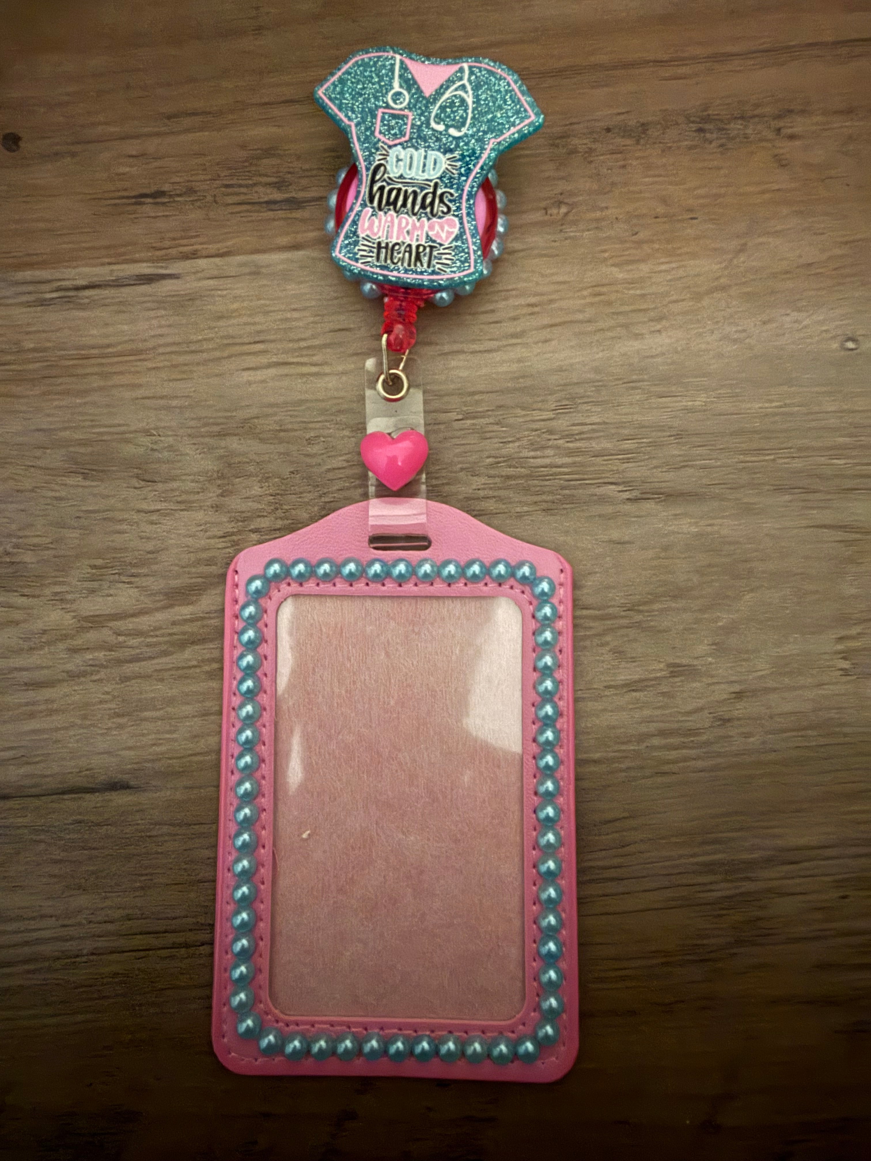 A pink ID badge holder decorated with light blue pearls, attached to a teal glittery scrub-top badge reel featuring the text "Cold Hands Warm Heart."