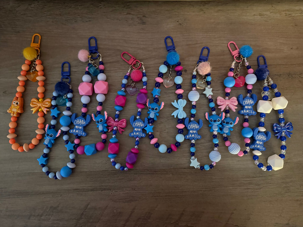 A variety of beaded lanyards and charms featuring an orange dog theme and several blue alien character themes with "Ohana means family" charms, decorated with pom-poms, bows, and stars.