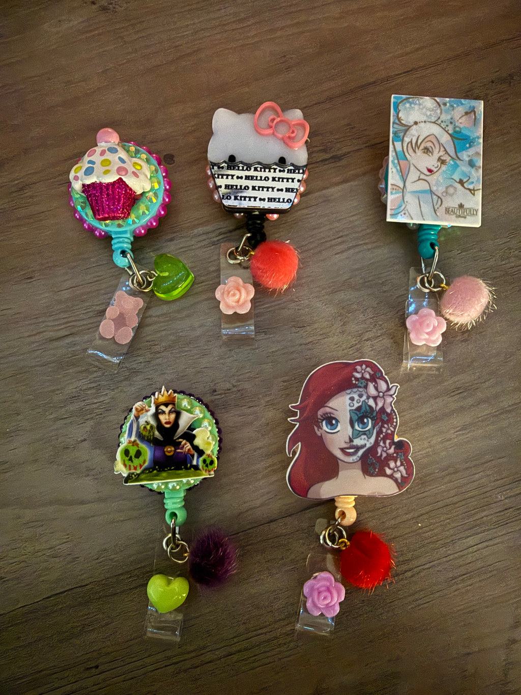 Five handcrafted decorative badge reels featuring a glittery cupcake, Hello Kitty with a pink bow, a retro Tinker Bell, the Evil Queen with poison apples, and a stylized Ariel with sugar skull face paint. Each reel is adorned with colorful charms, including pom-poms, flowers, and hearts.