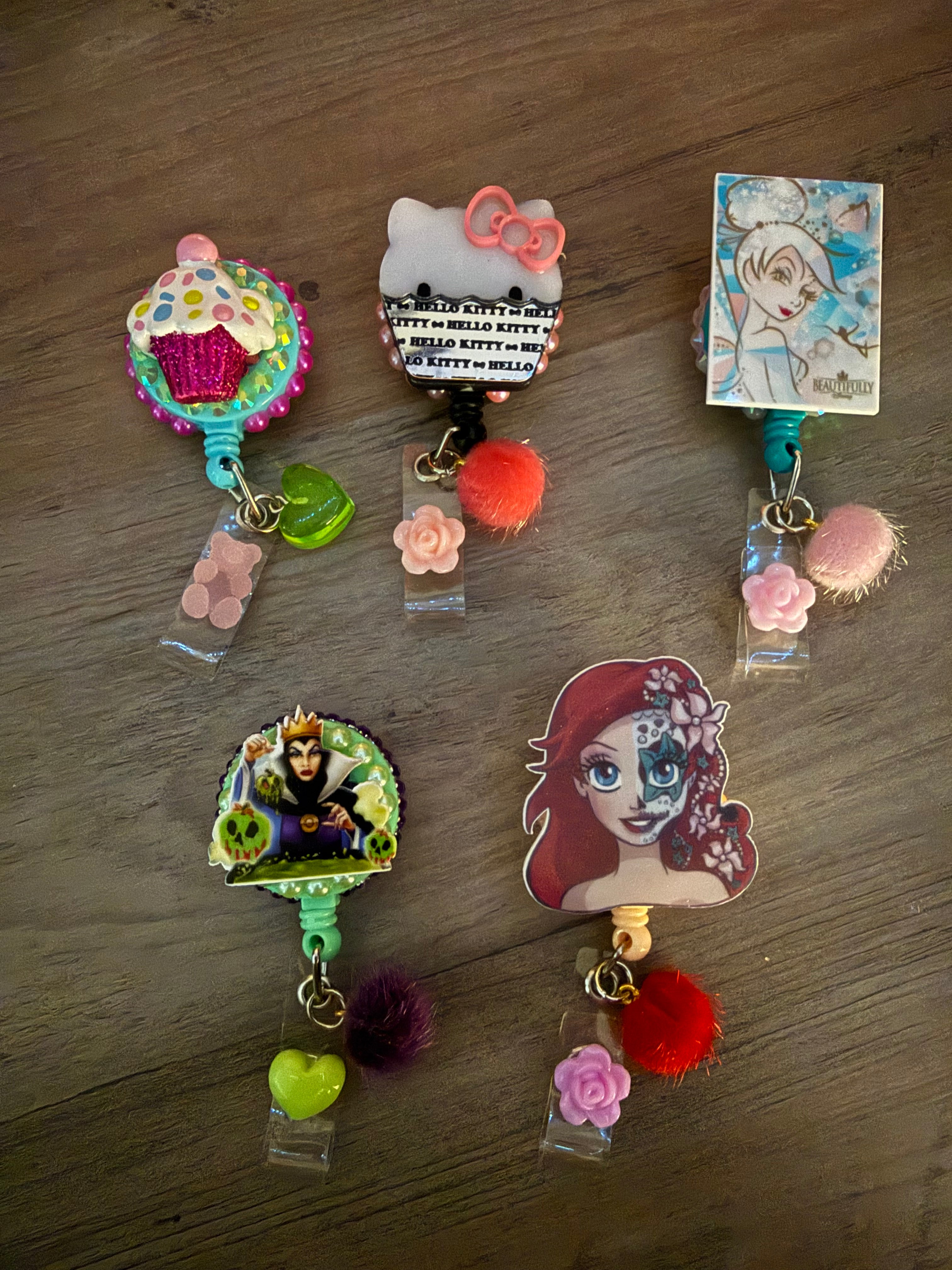 Five handcrafted decorative badge reels featuring a glittery cupcake, Hello Kitty with a pink bow, a retro Tinker Bell, the Evil Queen with poison apples, and a stylized Ariel with sugar skull face paint. Each reel is adorned with colorful charms, including pom-poms, flowers, and hearts.