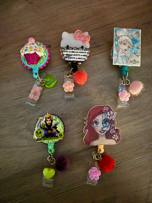 Five handcrafted decorative badge reels featuring a glittery cupcake, Hello Kitty with a pink bow, a retro Tinker Bell, the Evil Queen with poison apples, and a stylized Ariel with sugar skull face paint. Each reel is adorned with colorful charms, including pom-poms, flowers, and hearts.