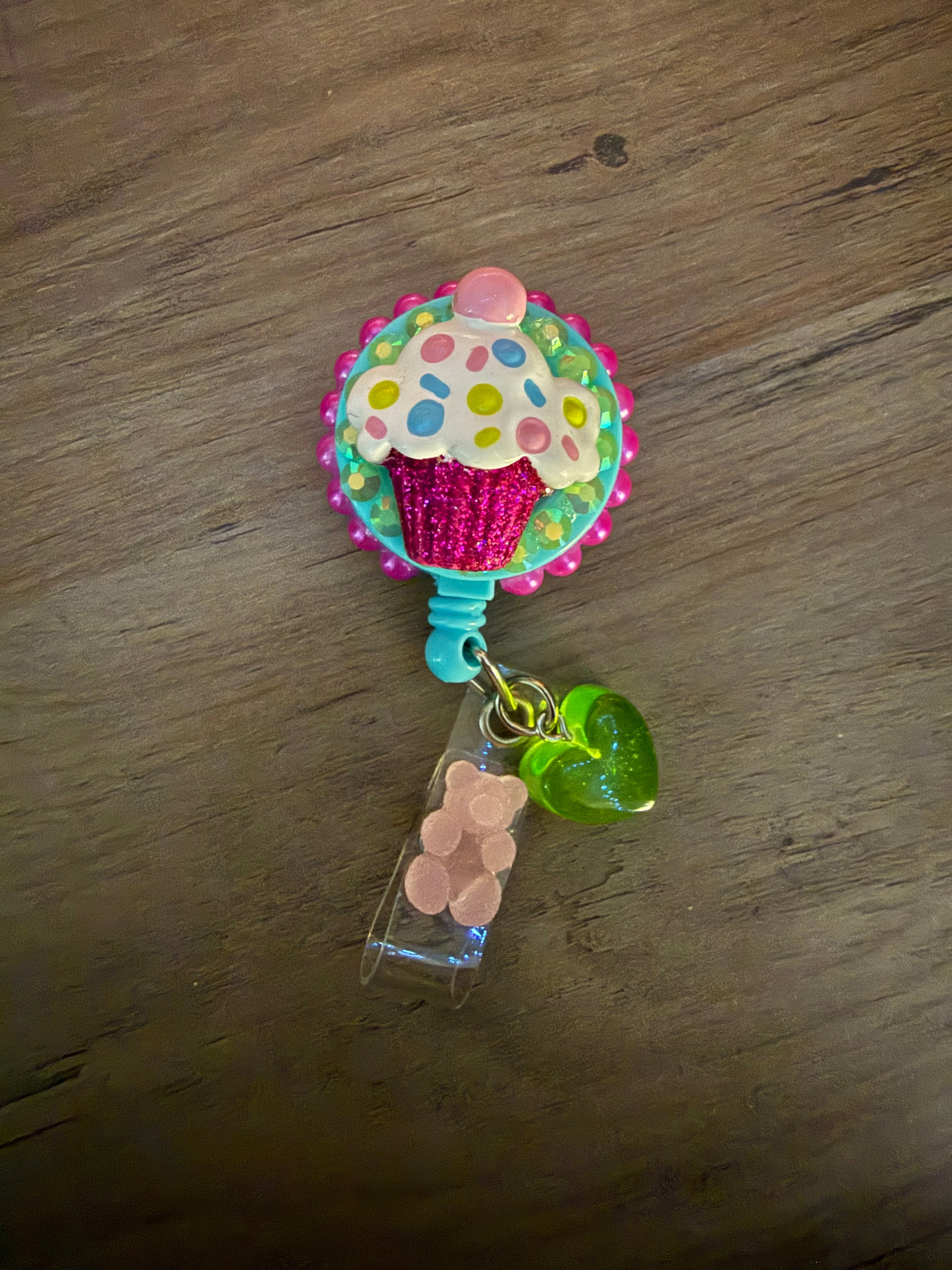A handcrafted retractable badge reel featuring a 3D pink glittery cupcake with colorful polka dots, set against a teal background with pink beaded trim. The reel includes a translucent green heart charm and a small pink gummy bear accent.