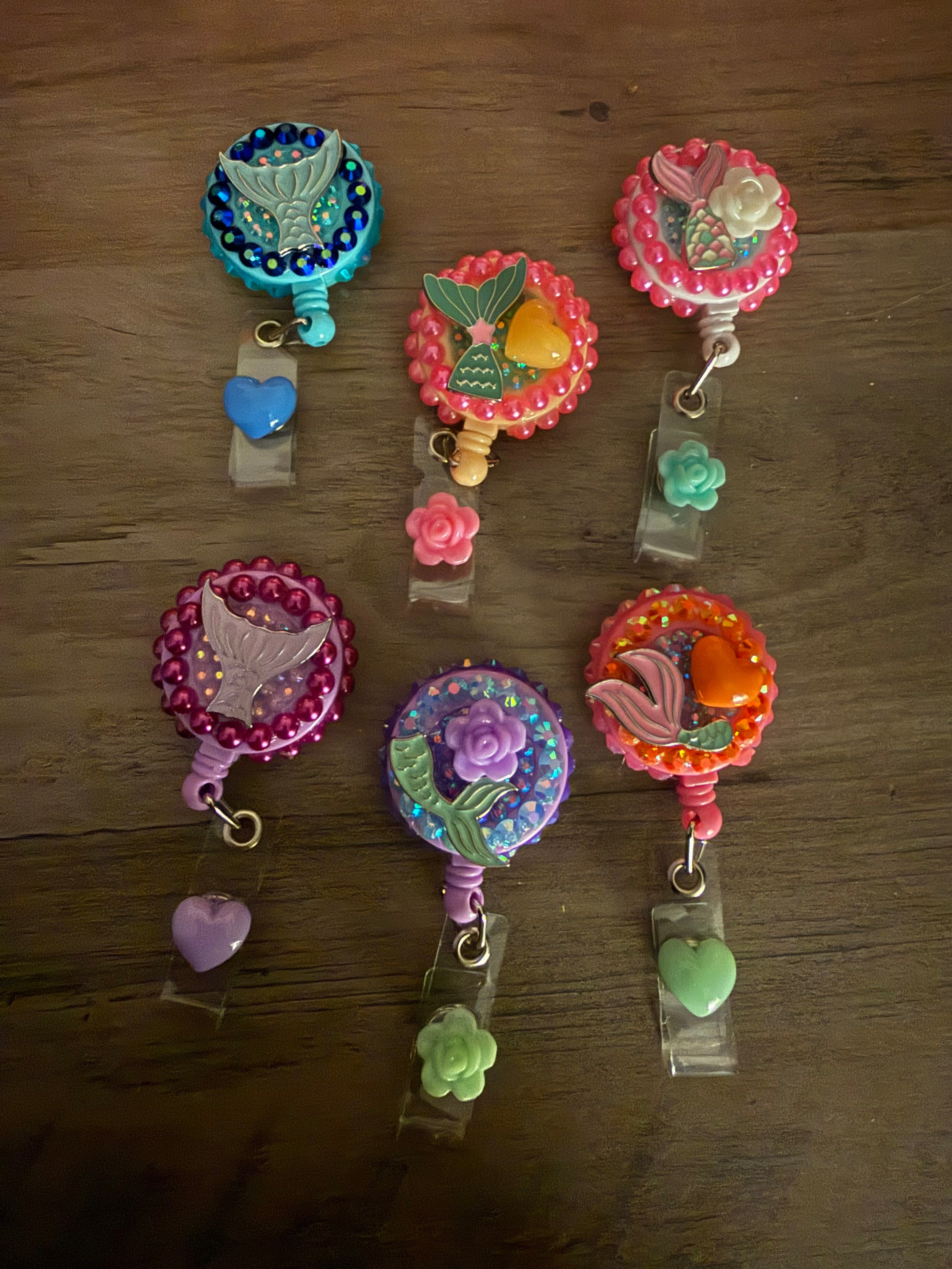 A collection of six handmade mermaid-themed badge reels in shades of blue, pink, purple, and orange, each decorated with glitter, pearls, and mermaid tail charms, featuring heart or flower dangles.