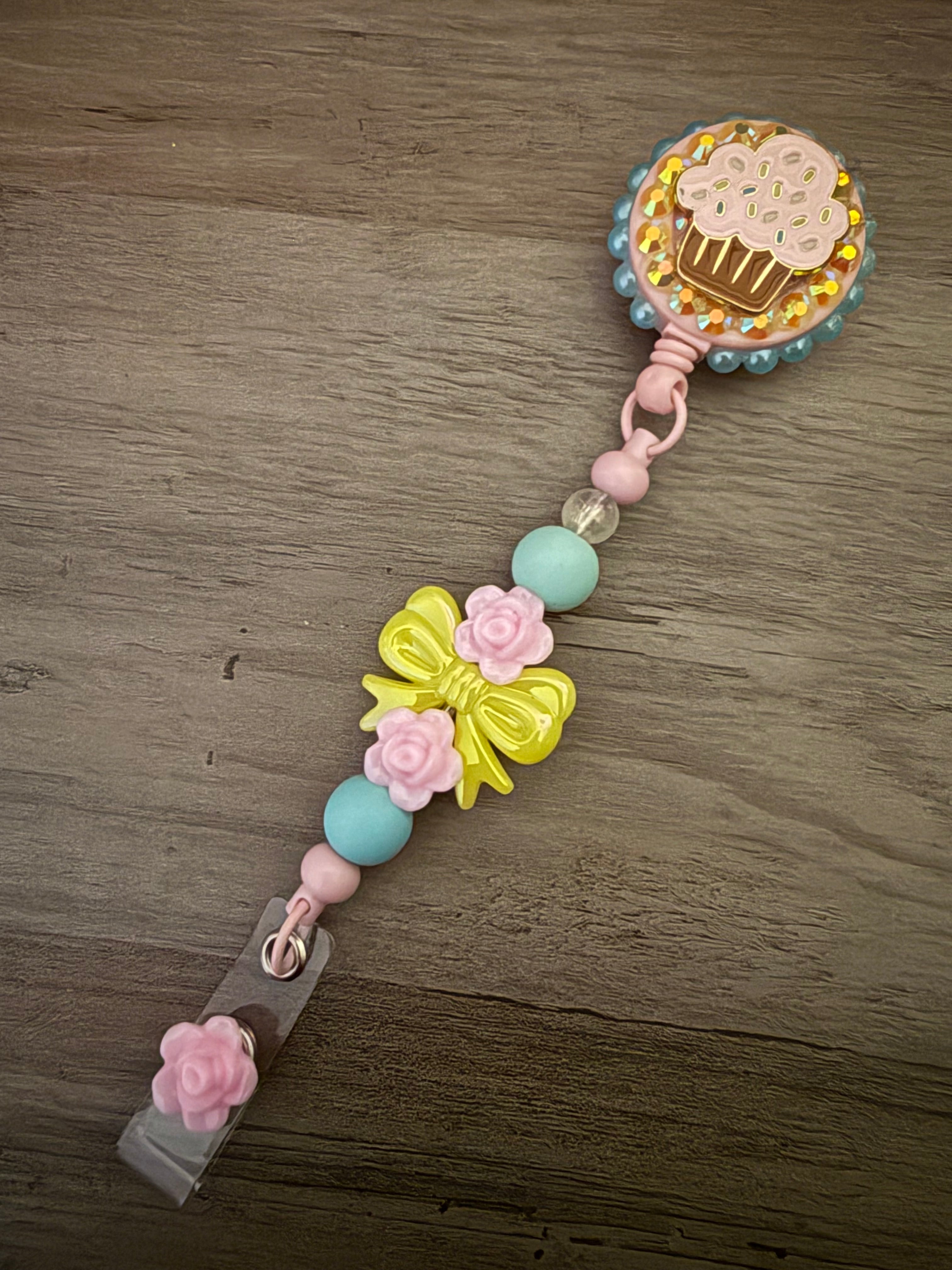 Handmade pink badge reel featuring a cupcake charm on a gold glittery base, accented by a decorative beaded lanyard with a yellow bow, pink roses, and a matching pink flower dangle.