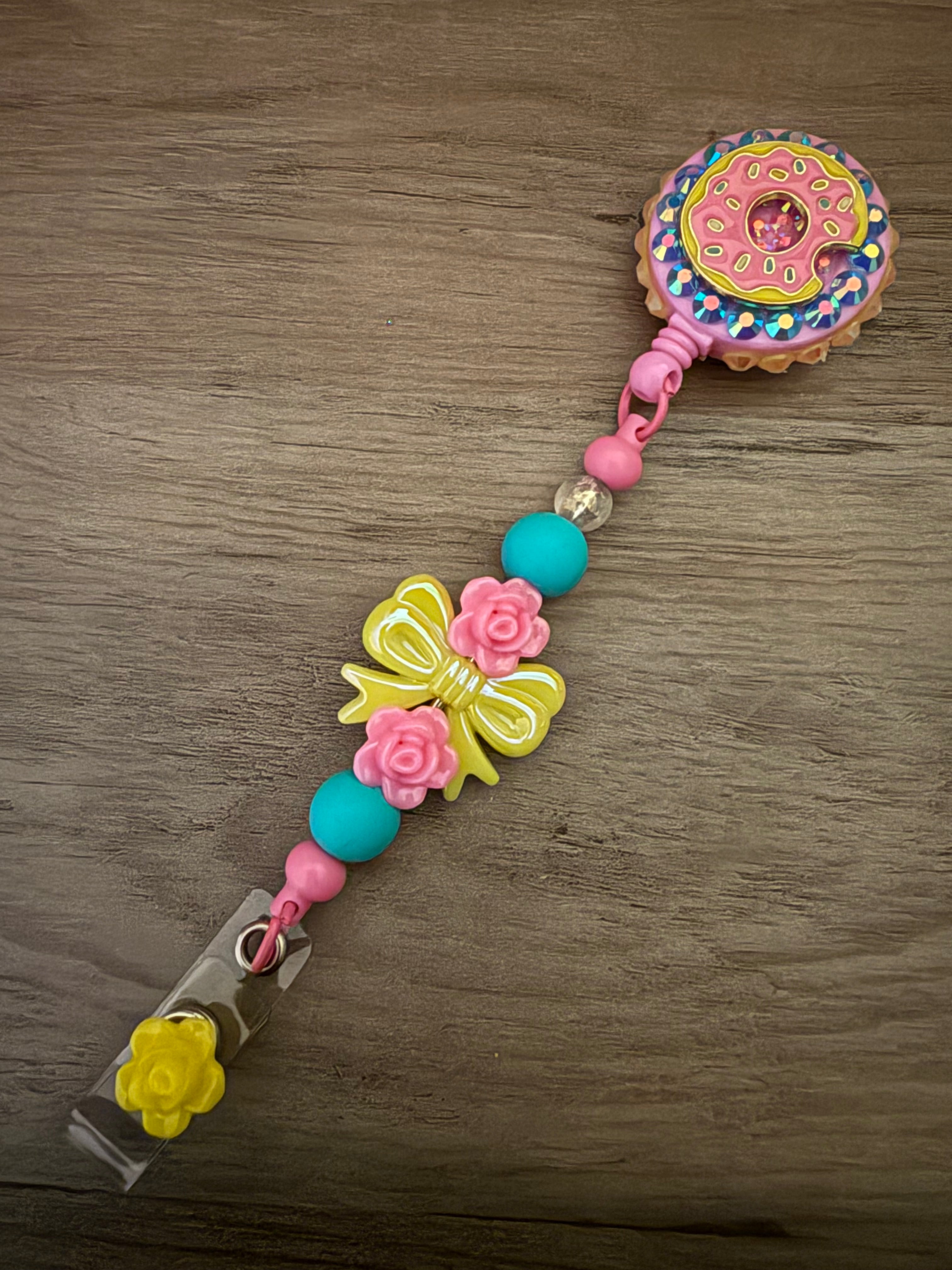 Handmade pink badge reel featuring a pink frosted donut charm on a purple glittery base, accented by a decorative beaded lanyard with a yellow bow, pink roses, and a yellow flower dangle.