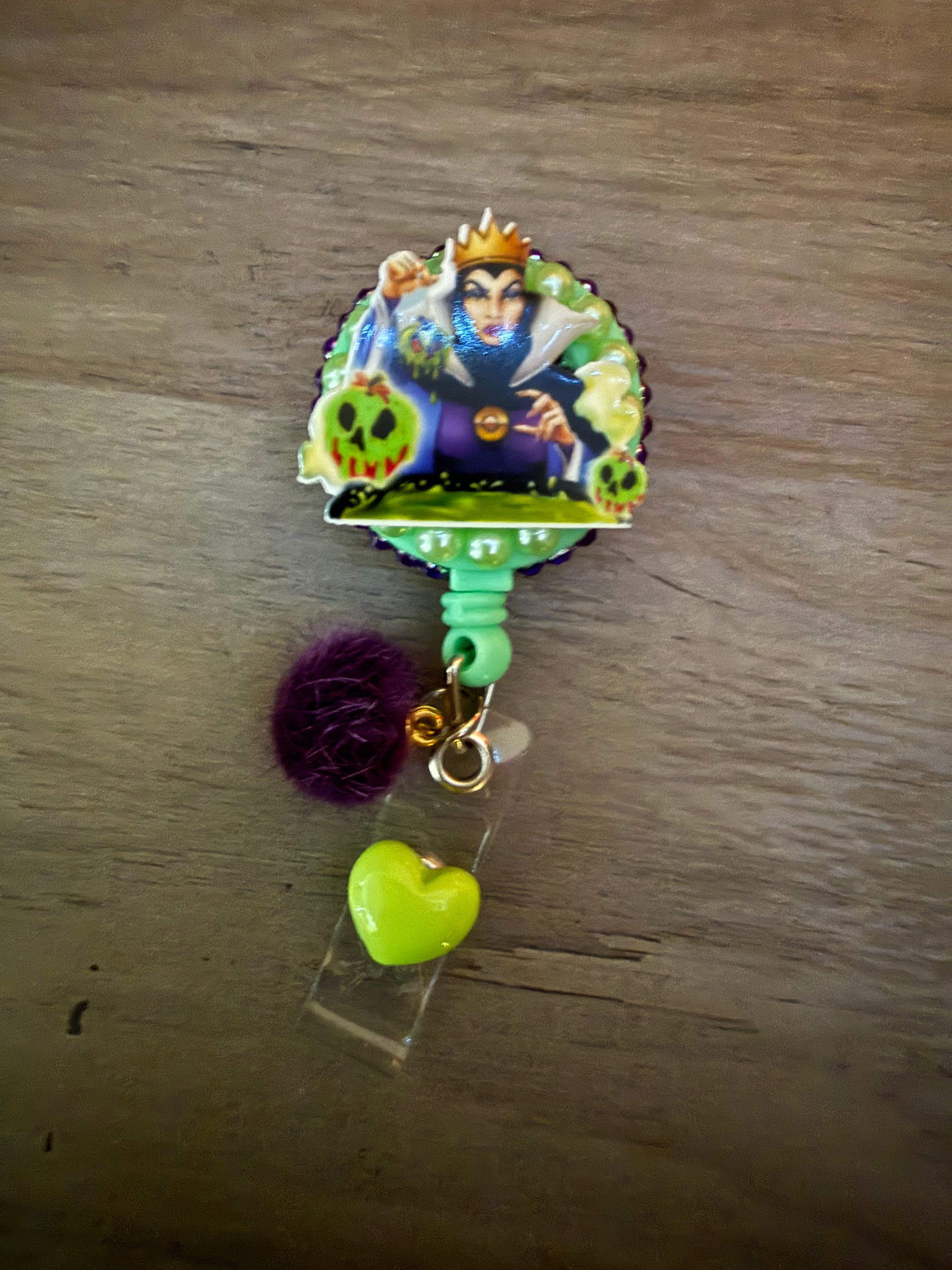 A handcrafted retractable badge reel featuring the Evil Queen holding a poison apple over a cauldron. The design is framed with light green beads and dark purple trim, accented with a dark purple fuzzy pom-pom and a lime green heart charm.