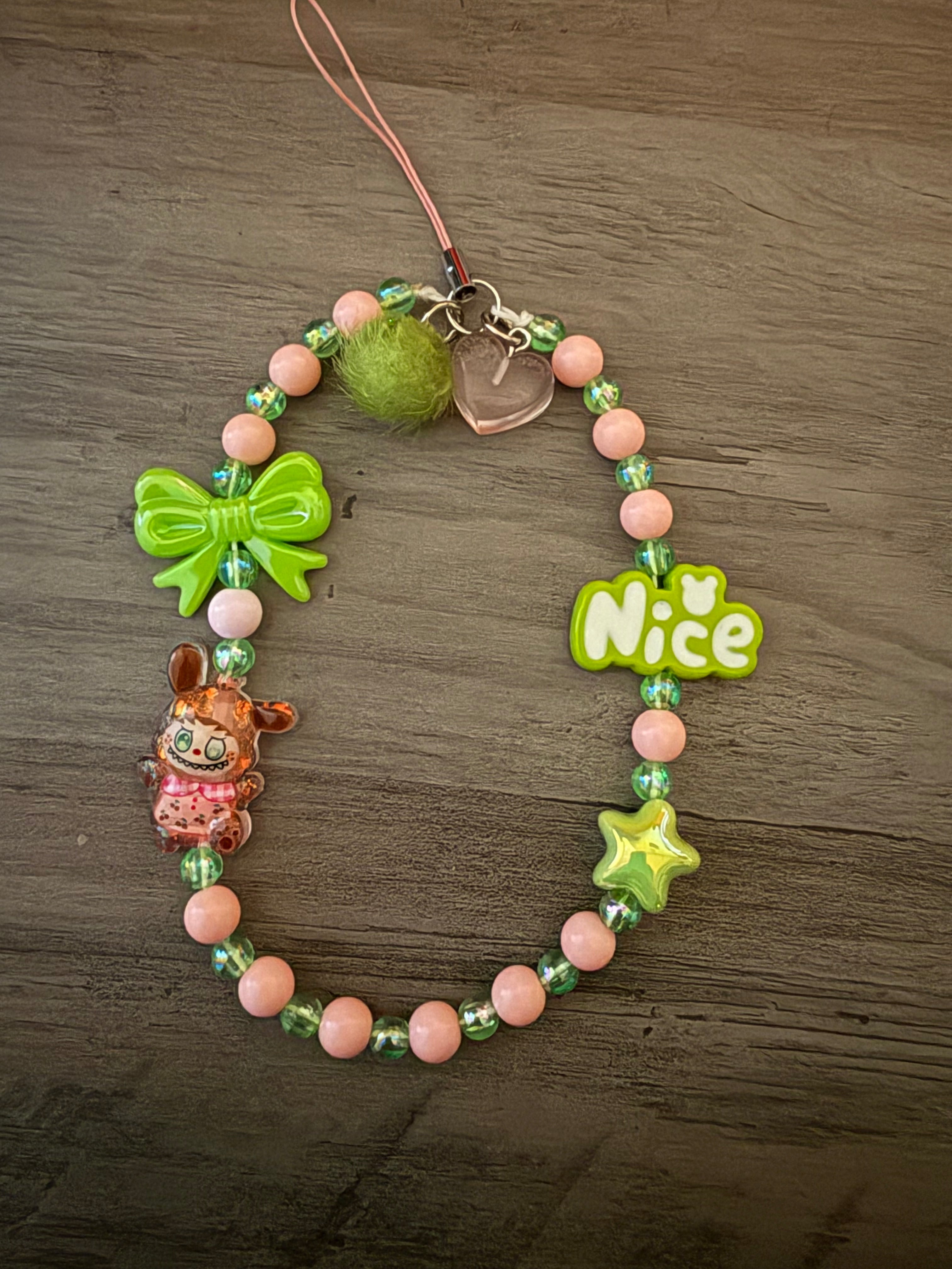 A beaded phone charm featuring lime green and peach beads, a green bow, a green star, a "Nice" word charm, and a small brown monster charm on a wooden background.