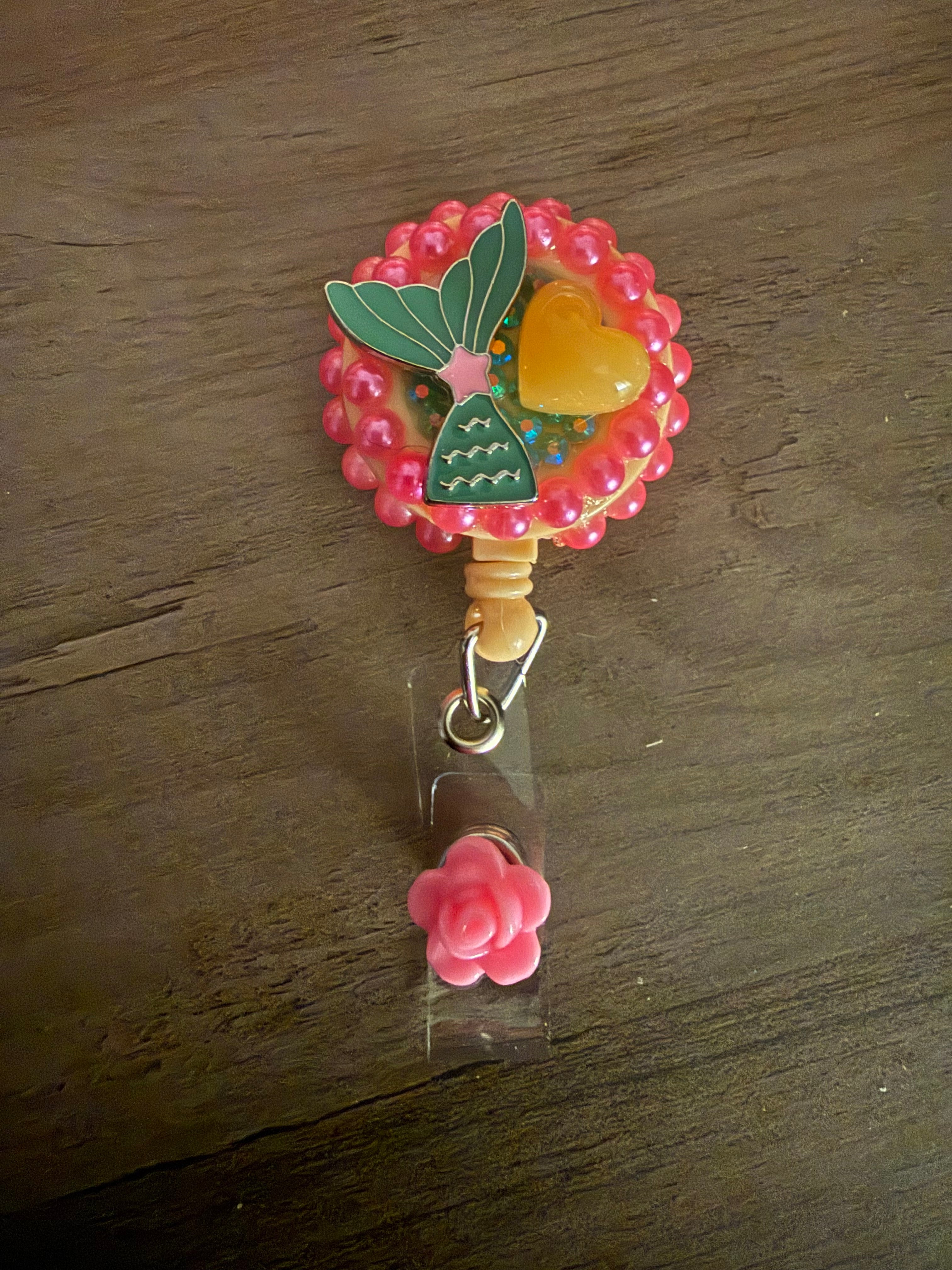 Handmade peach badge reel decorated with pink pearls, a green mermaid tail, and a yellow heart charm, finished with a pink rose dangle.