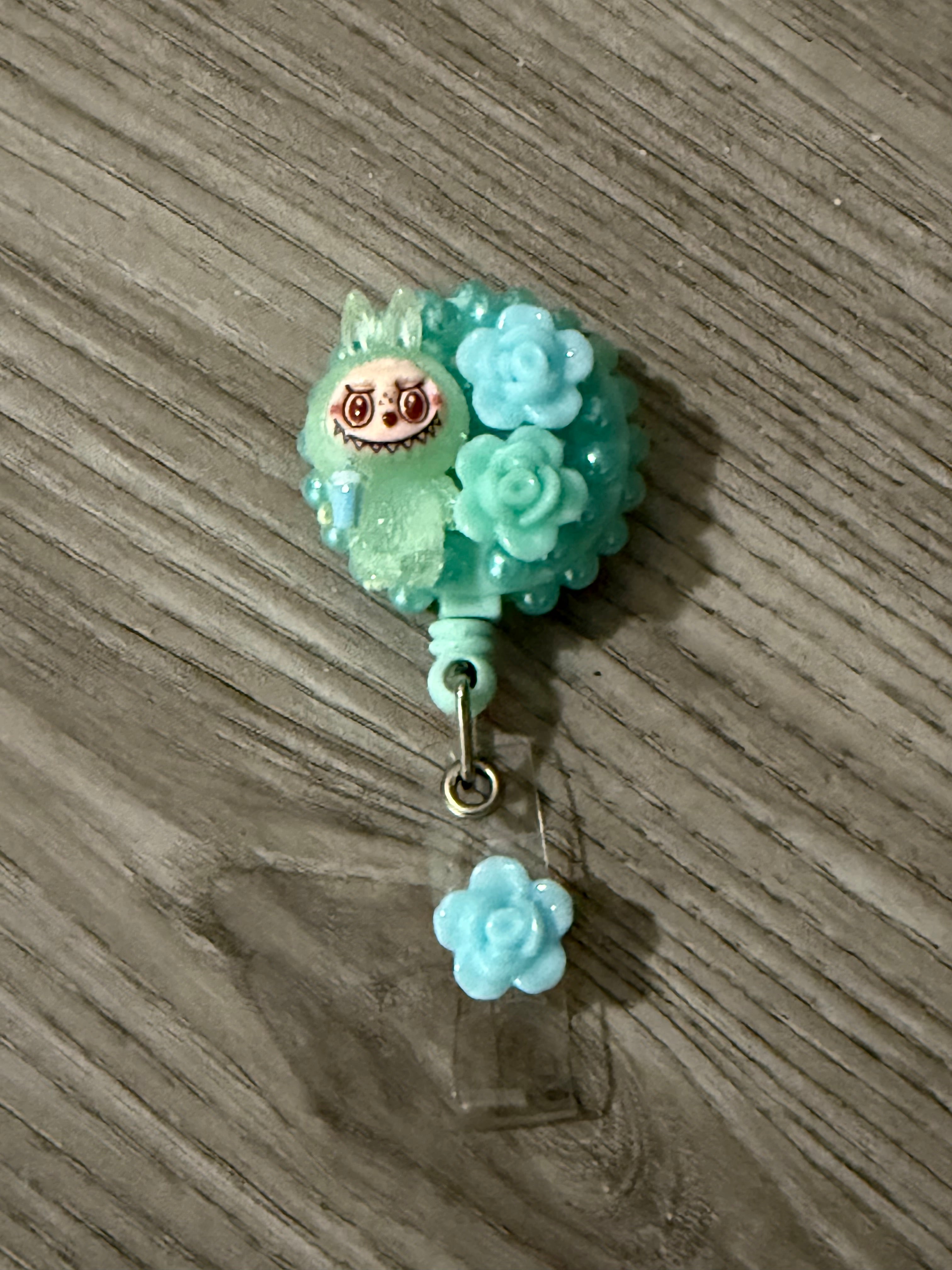 Cute badge reel featuring a whimsical green monster with bunny ears, surrounded by blue roses and beads, with a small blue flower charm attached.