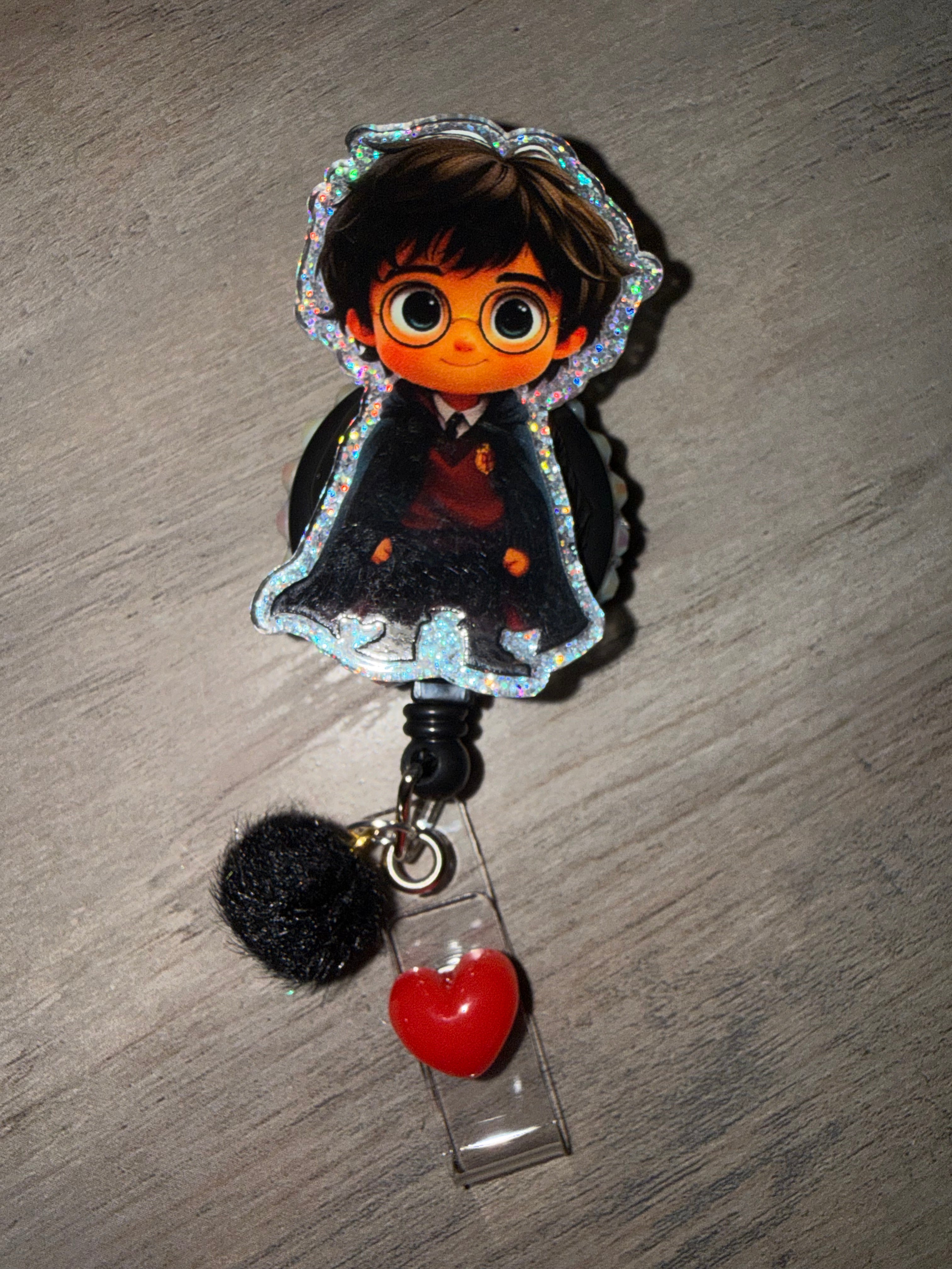 A glittery badge reel featuring a chibi-style Harry Potter character. The acrylic charm is bordered with silver glitter and attached to a black retractable cord, decorated with a small black pom-pom and a bright red heart charm hanging from the clear strap.