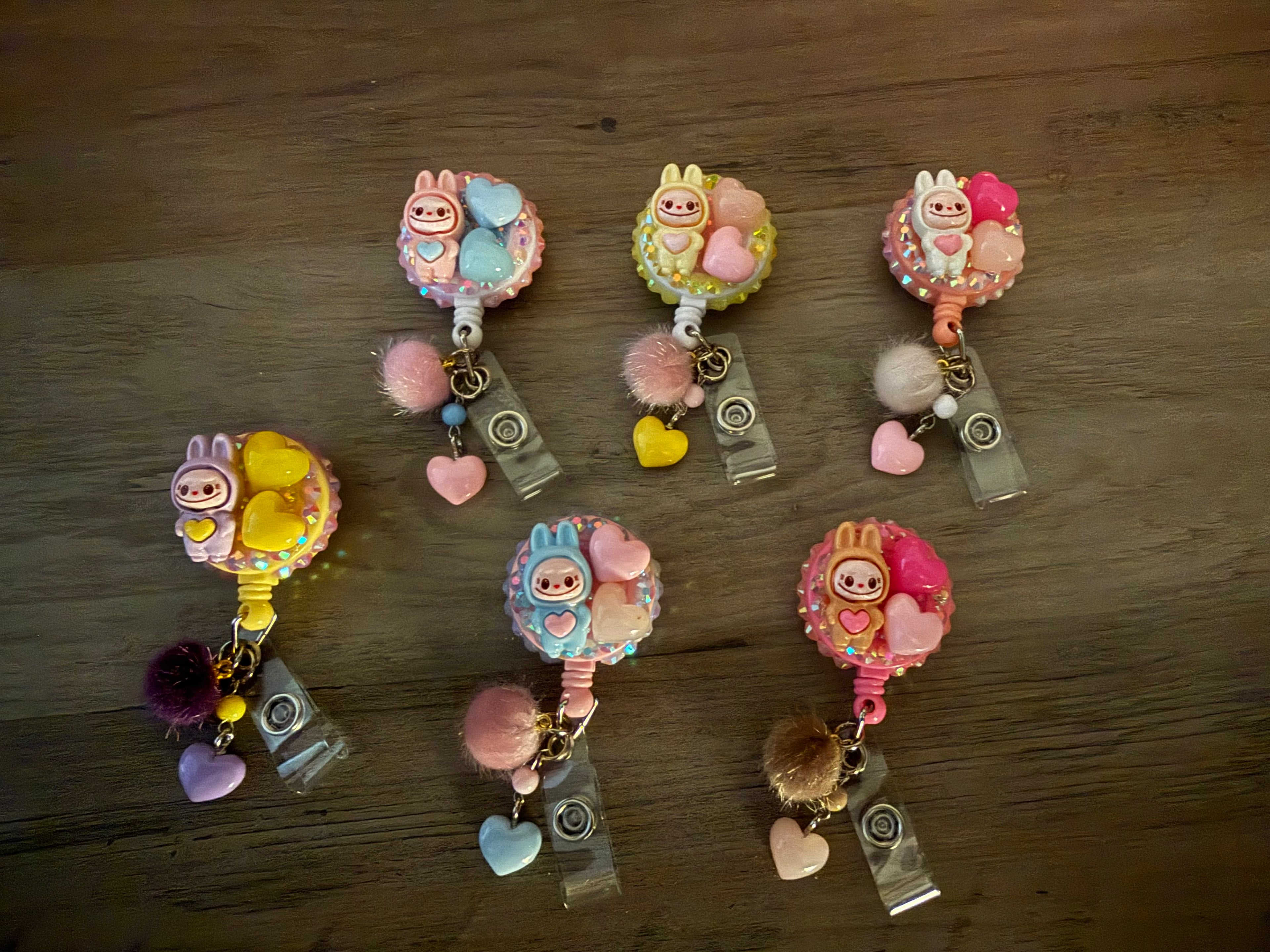 Six colorful, handcrafted retractable badge reels in pastel shades, each decorated with a monster figure, heart charms, rhinestones, and a small pom-pom.