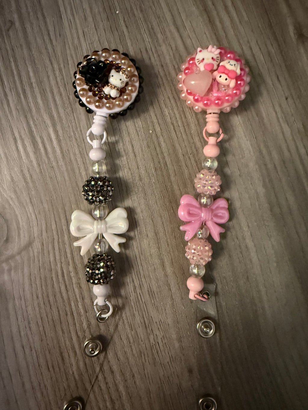 Two decoden-style retractable badge reels featuring Sanrio characters. The left reel is black, white, and bronze with a Hello Kitty figure, black rose, and white bow. The right reel is monochromatic pink with Hello Kitty and My Melody figures, a heart, and a pink bow. Both are accented with rhinestones and decorative beads.