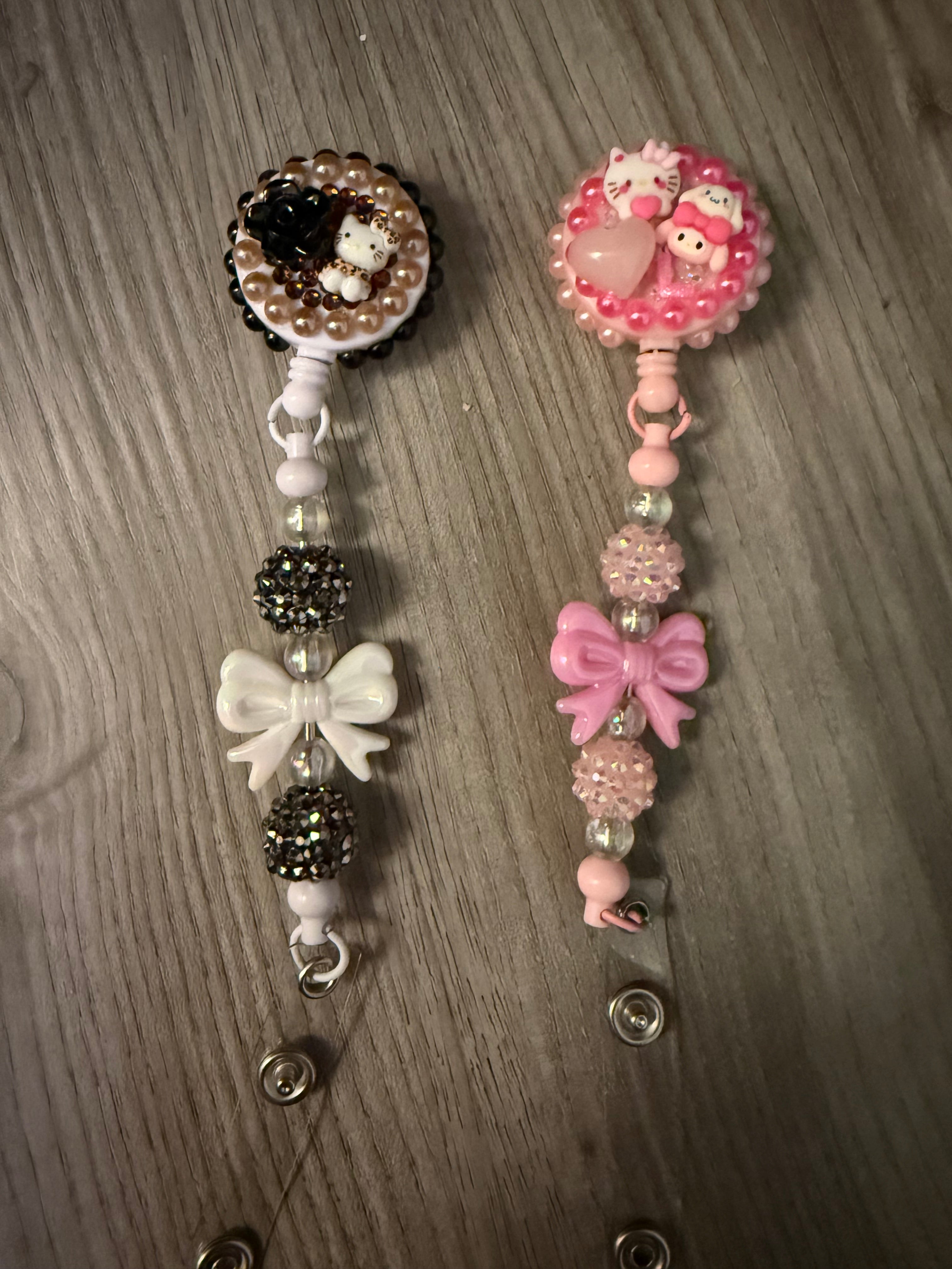 Two decoden-style retractable badge reels featuring Sanrio characters. The left reel is black, white, and bronze with a Hello Kitty figure, black rose, and white bow. The right reel is monochromatic pink with Hello Kitty and My Melody figures, a heart, and a pink bow. Both are accented with rhinestones and decorative beads.