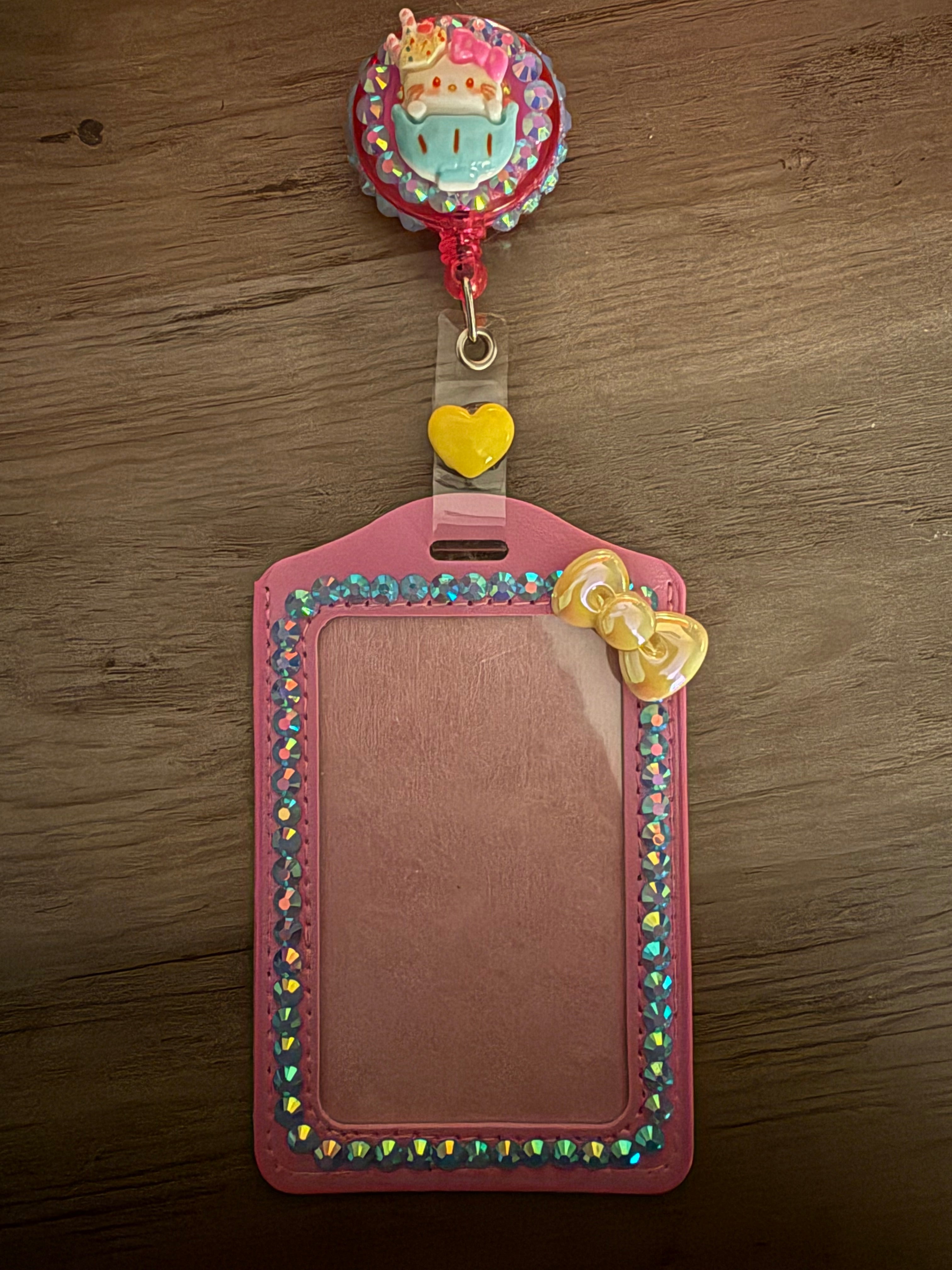 A pink, rhinestone-embellished ID card holder with a matching retractable badge reel. The reel features a Hello Kitty character charm sitting in a blue teacup against a glittering background, and the holder is lined with iridescent blue gems and a small yellow pearlescent bow.