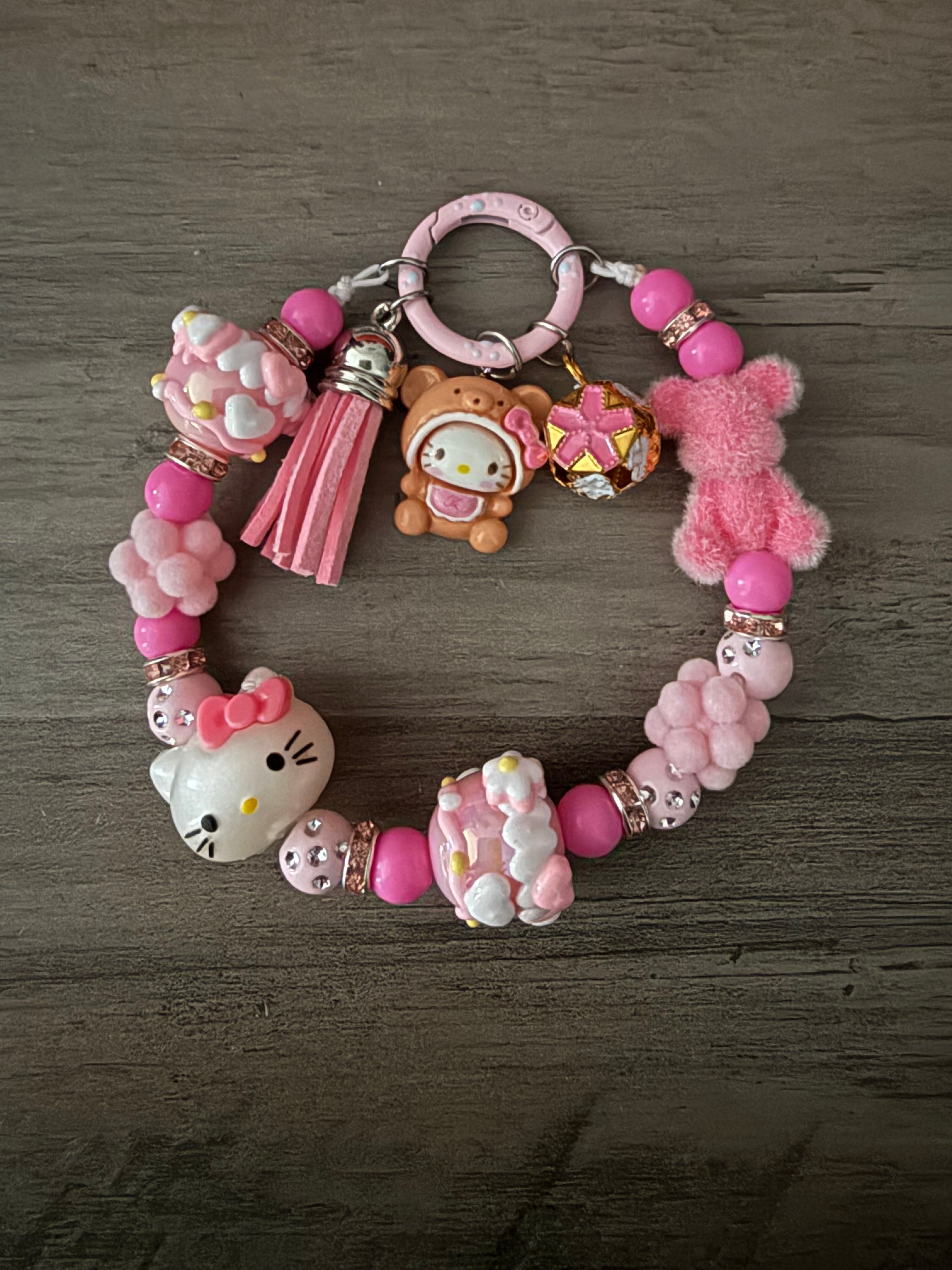 A pink-themed beaded charm featuring Hello Kitty figures, a fuzzy pink bear, and a pink tassel. The beads include hot pink and light pink rounds, rose gold rhinestone spacers, and pom-pom accents, all attached to a pastel pink circular clasp.