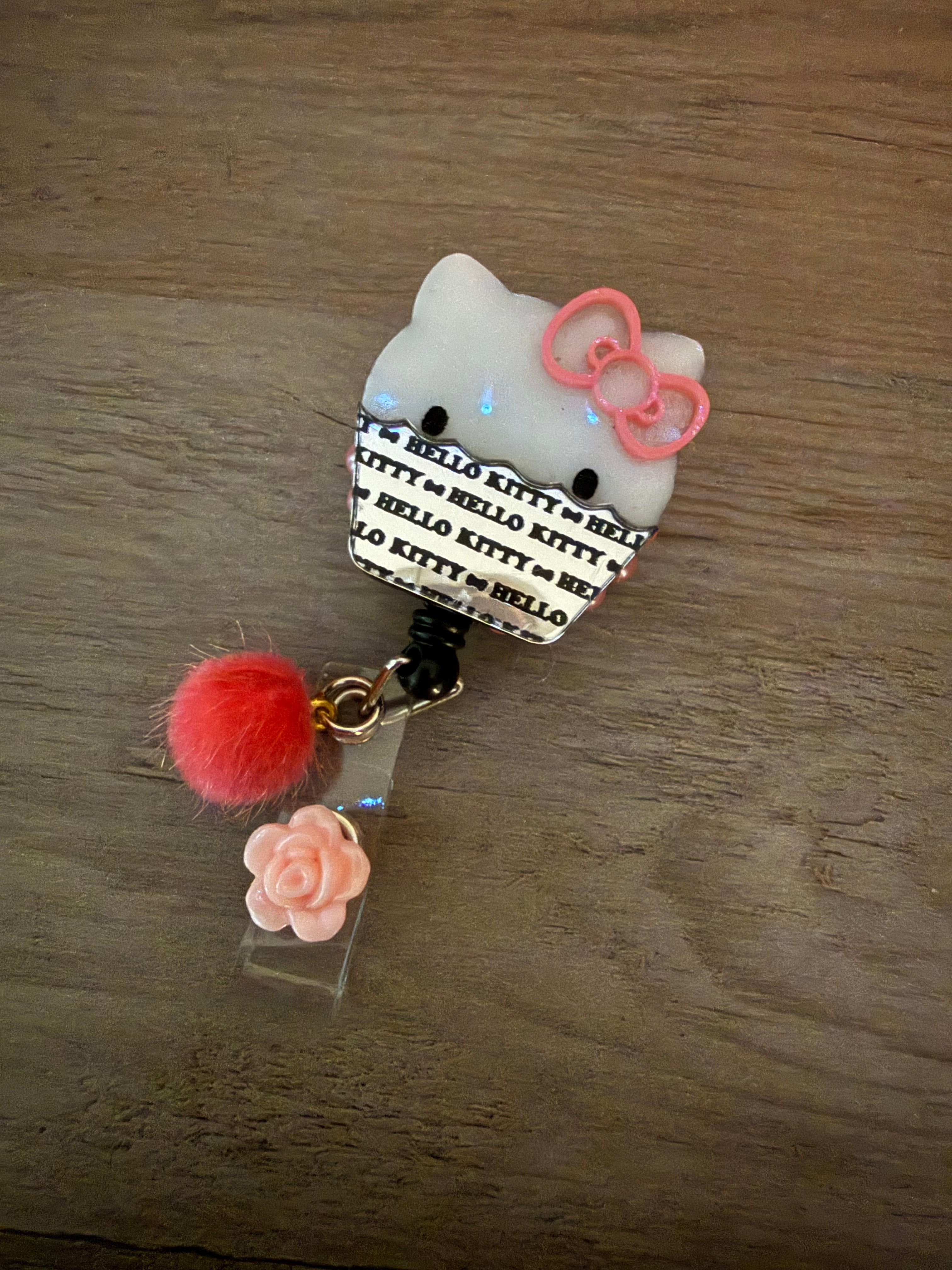 A handcrafted retractable badge reel featuring a Hello Kitty cupcake design with a pink bow and sparkly white icing. The reel is embellished with a small red fuzzy pom-pom and a light pink rose charm.