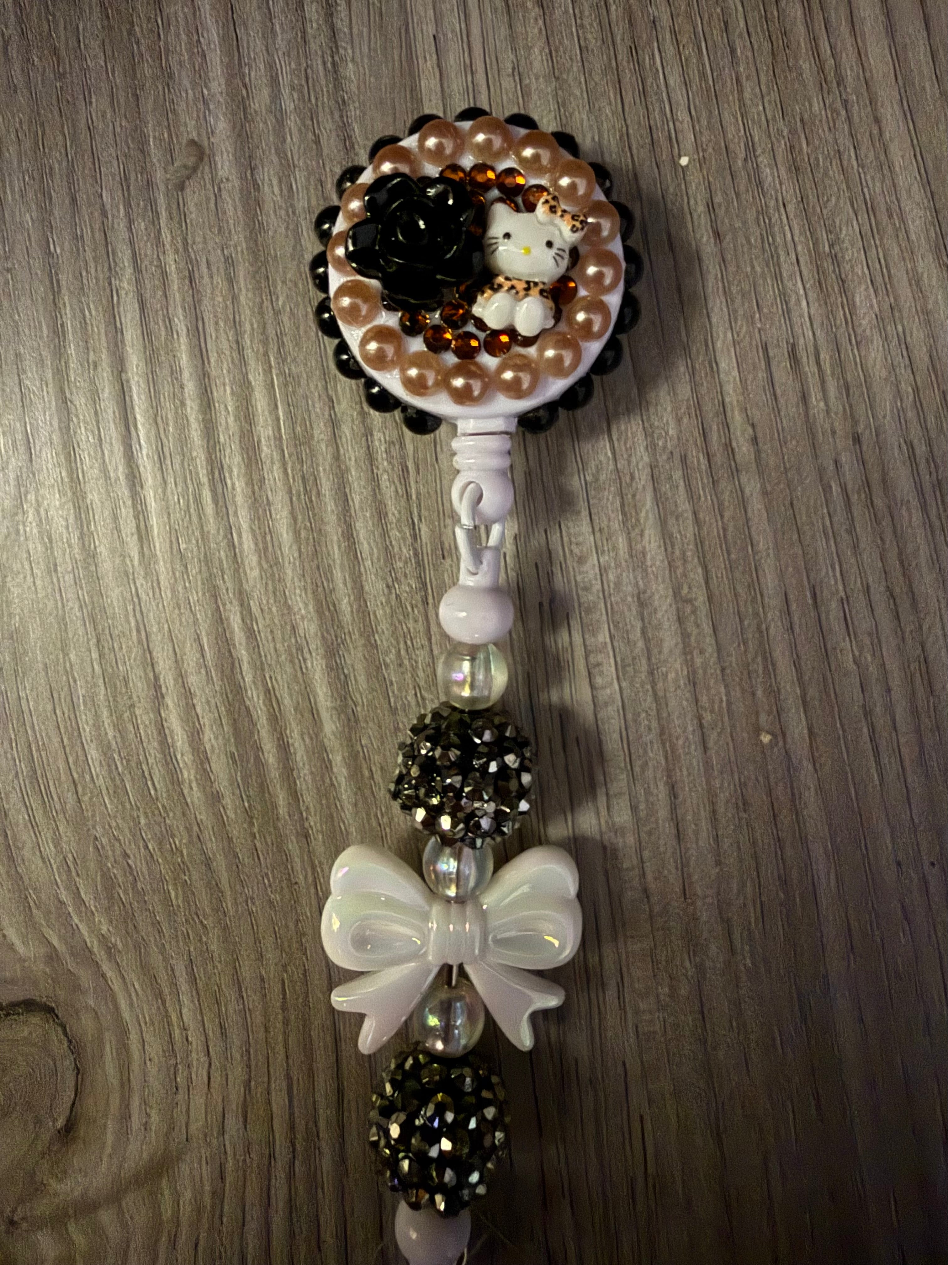 Handmade decoden retractable badge reel featuring a Hello Kitty figure in leopard print, a black rose, and bronze pearls. The beaded lead includes a white iridescent bow and sparkling disco ball beads.