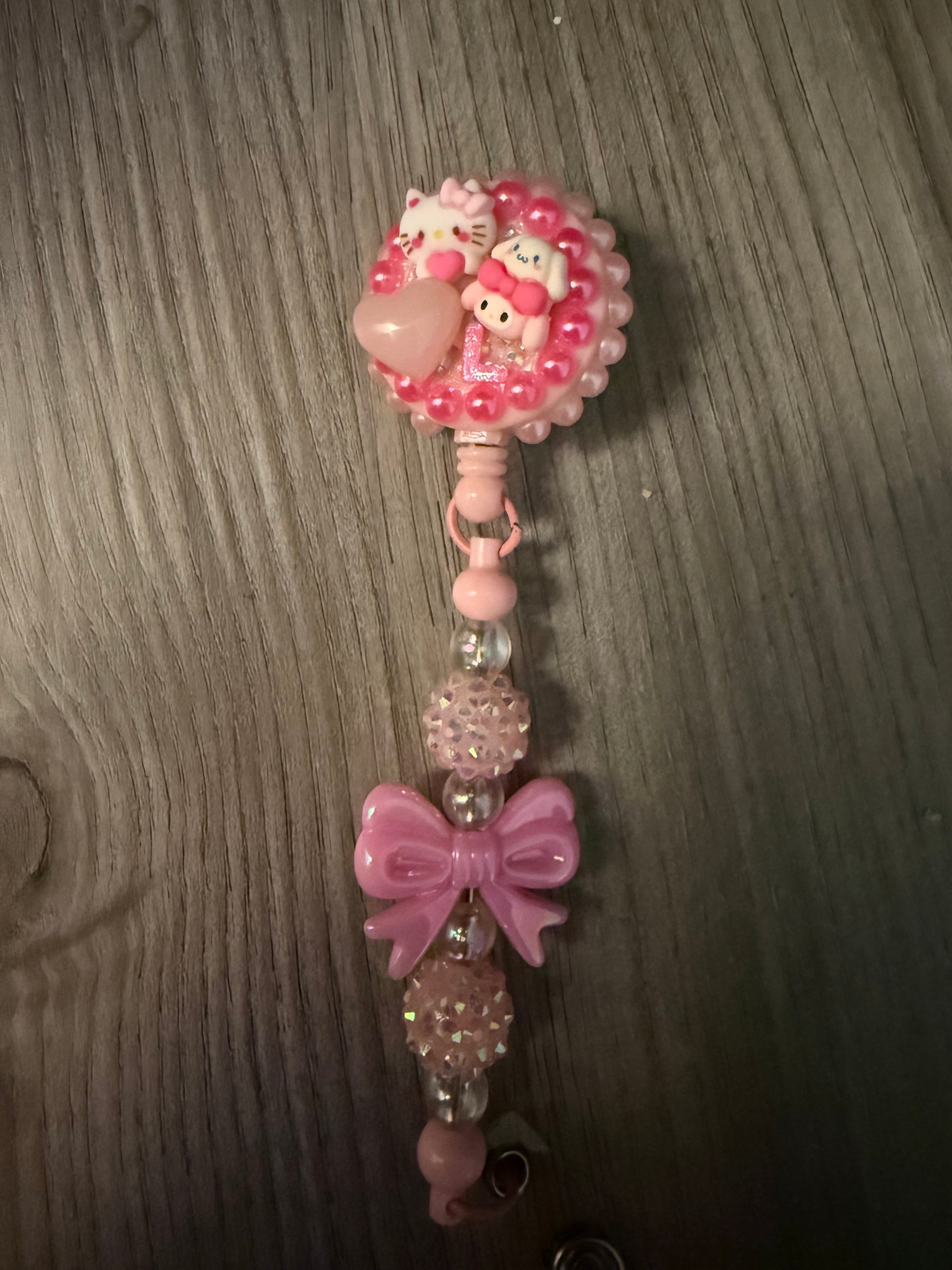 Handmade pink decoden retractable badge reel decorated with Hello Kitty, My Melody, and Cinnamoroll figures, a translucent heart, and pink pearls. The matching beaded lead features a large pink bow and glittery disco ball beads.