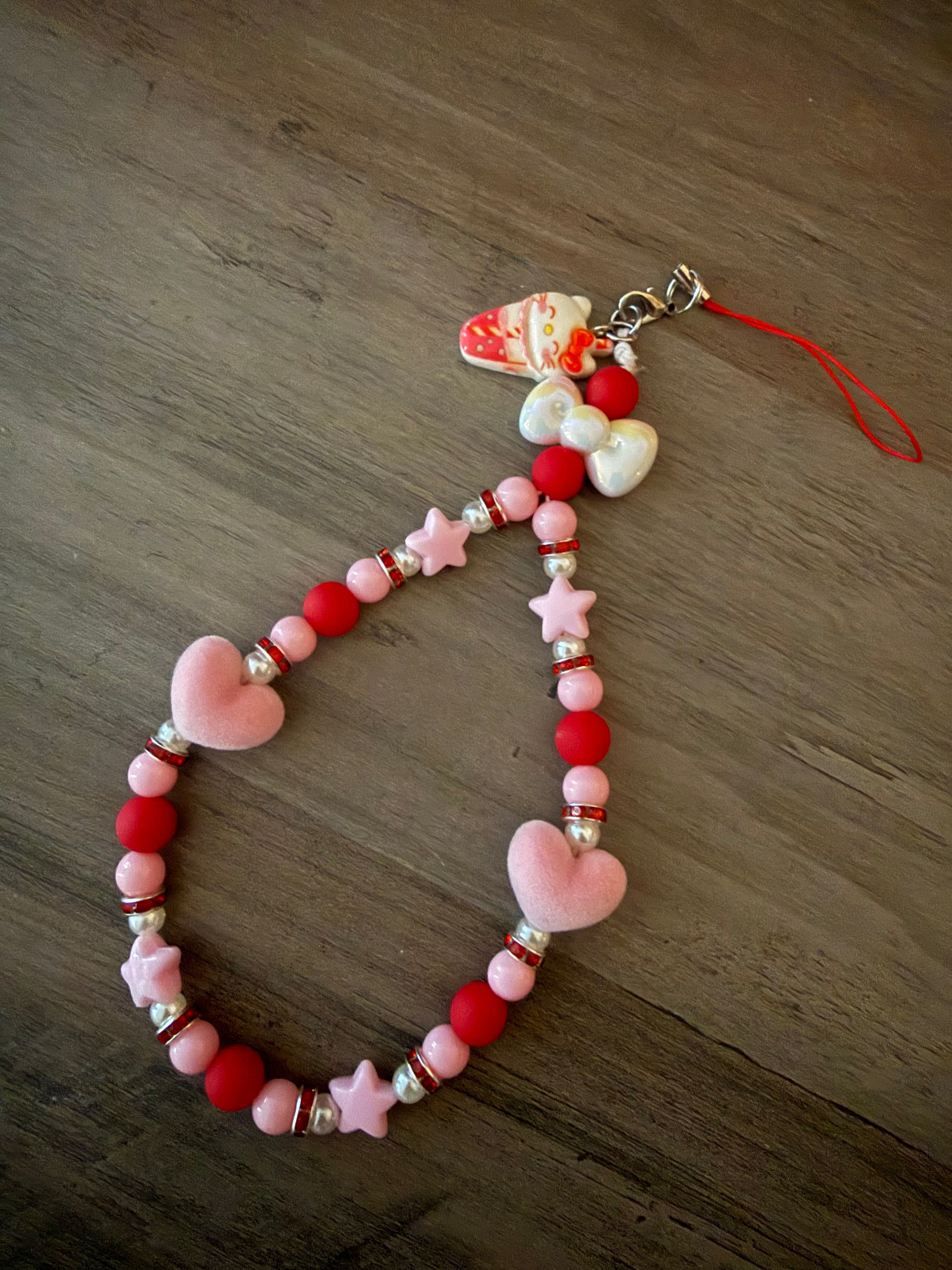 A handmade beaded phone wristlet featuring a mix of red and pink matte beads, sparkly spacers, and pink star and heart charms, finished with a white bow and a Hello Kitty strawberry drink charm.