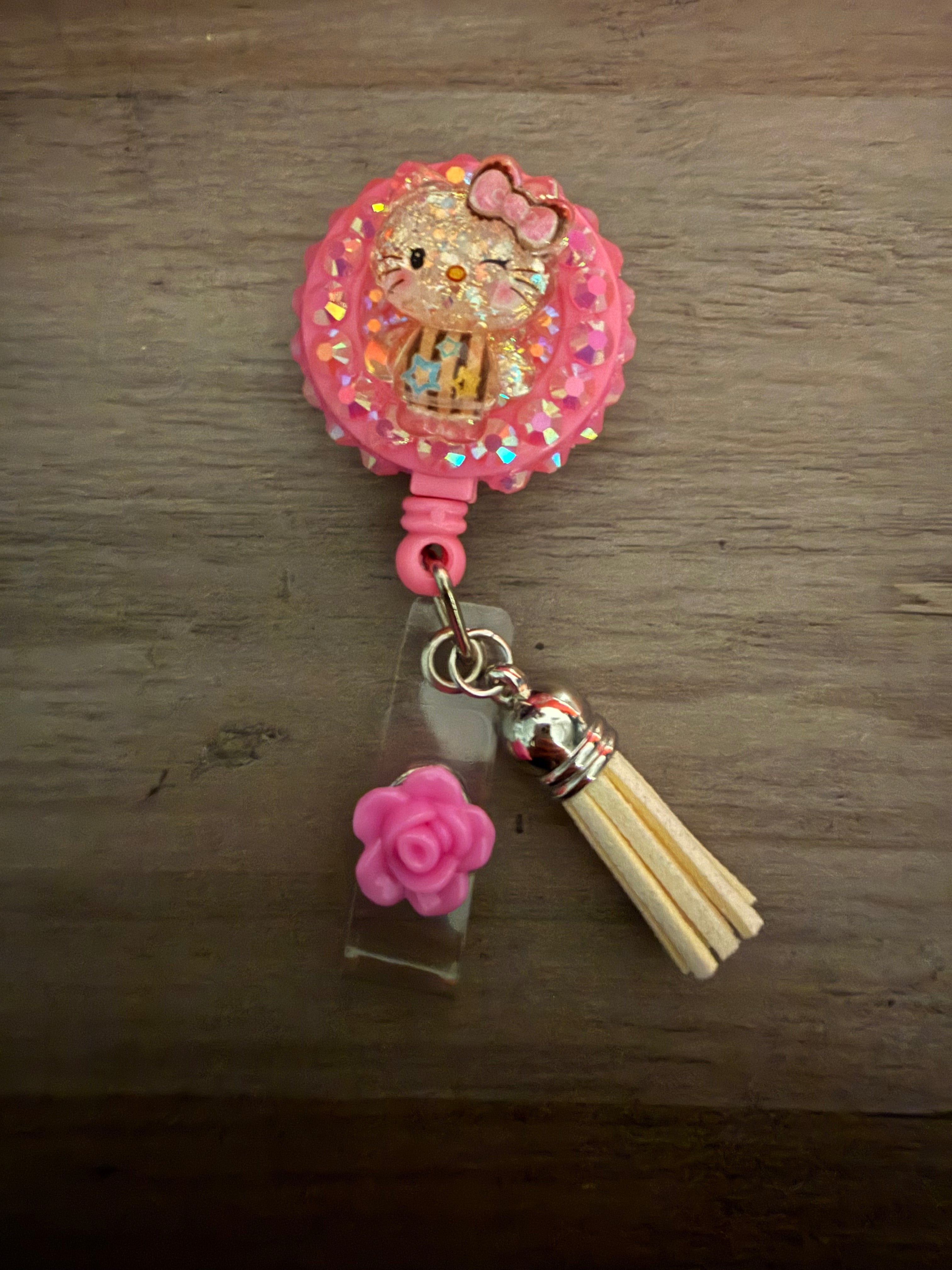 A pink decoden retractable badge reel featuring a glittery Hello Kitty charm, pink rhinestones, a cream-colored tassel, and a small pink rose accent.