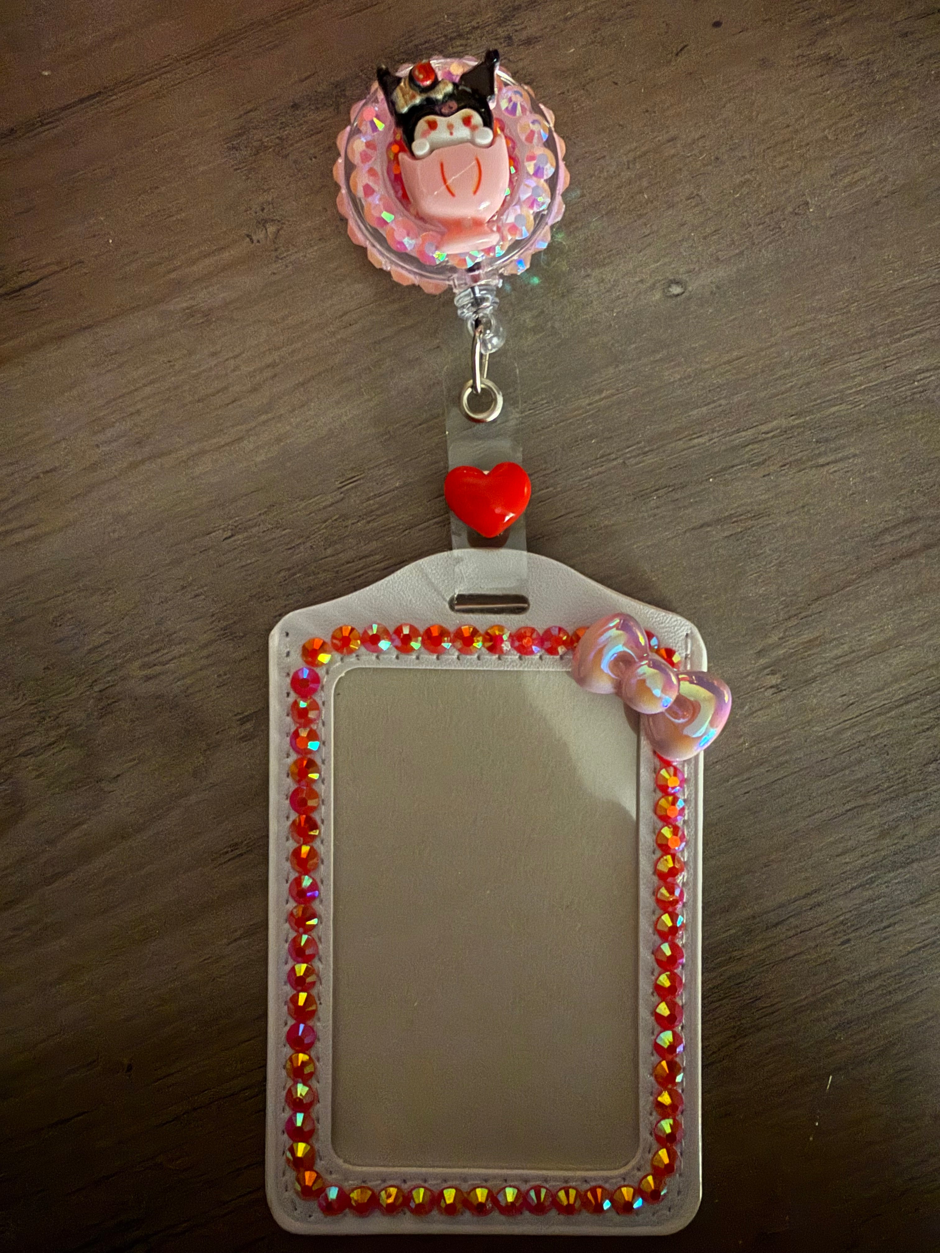 A white, rhinestone-embellished ID card holder with a matching retractable badge reel. The reel features a Kuromi character charm sitting in a pink sundae cup against a sparkling background, and the holder is lined with iridescent orange gems and a small pink pearlescent bow.