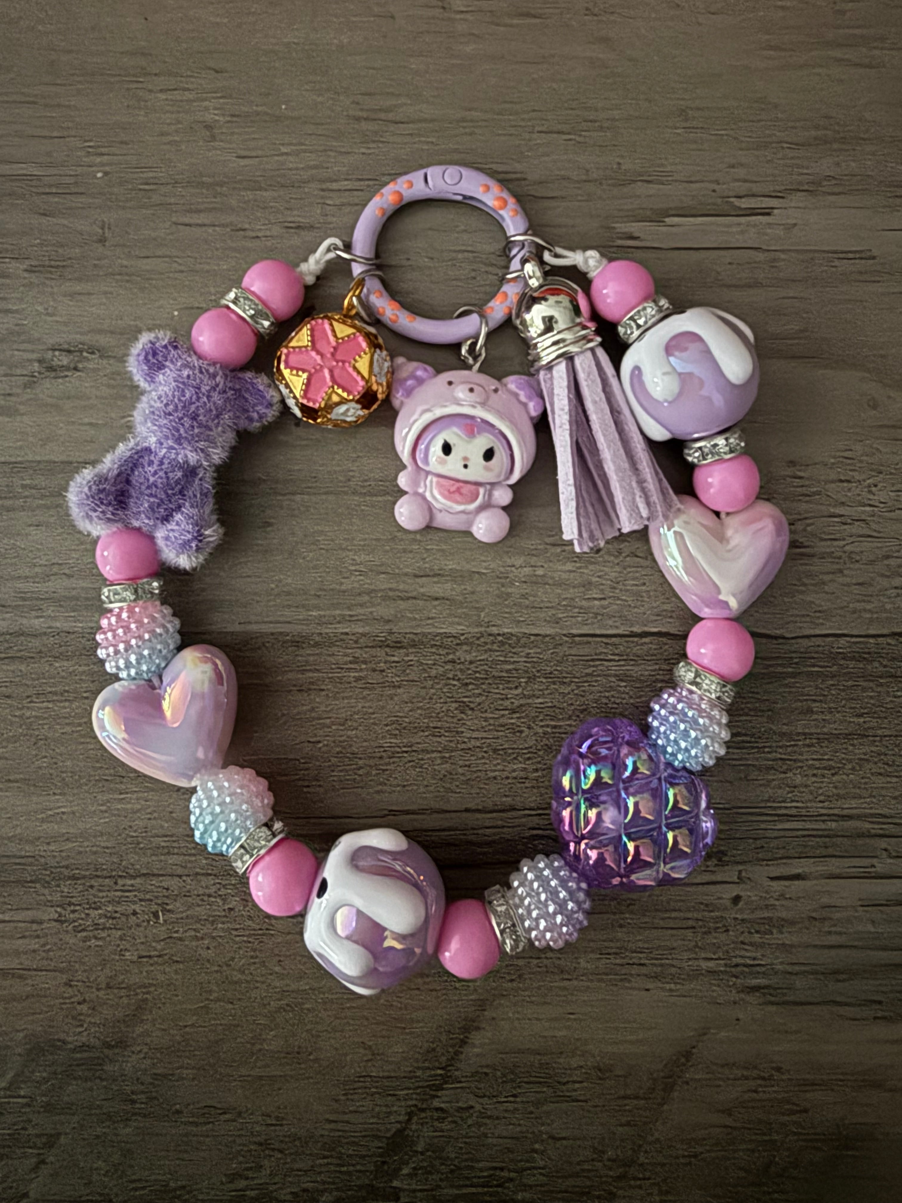 A purple and pink beaded charm featuring Kuromi figure, a fuzzy purple bear, and a purple tassel. The beads include pink rounds, iridescent purple hearts, textured "berry" beads, and silver rhinestone spacers, all attached to a lilac circular clasp.