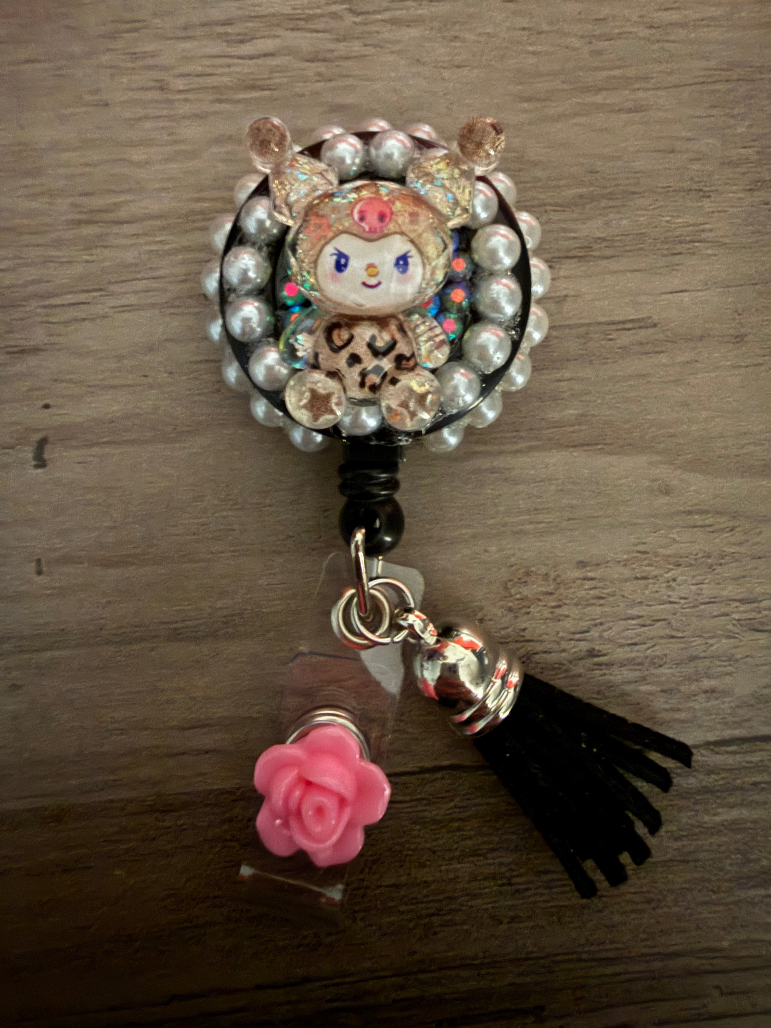 A decoden retractable badge reel featuring a glittery Kuromi character in leopard print, bordered by white pearls and accented with a black tassel and pink rose charm.