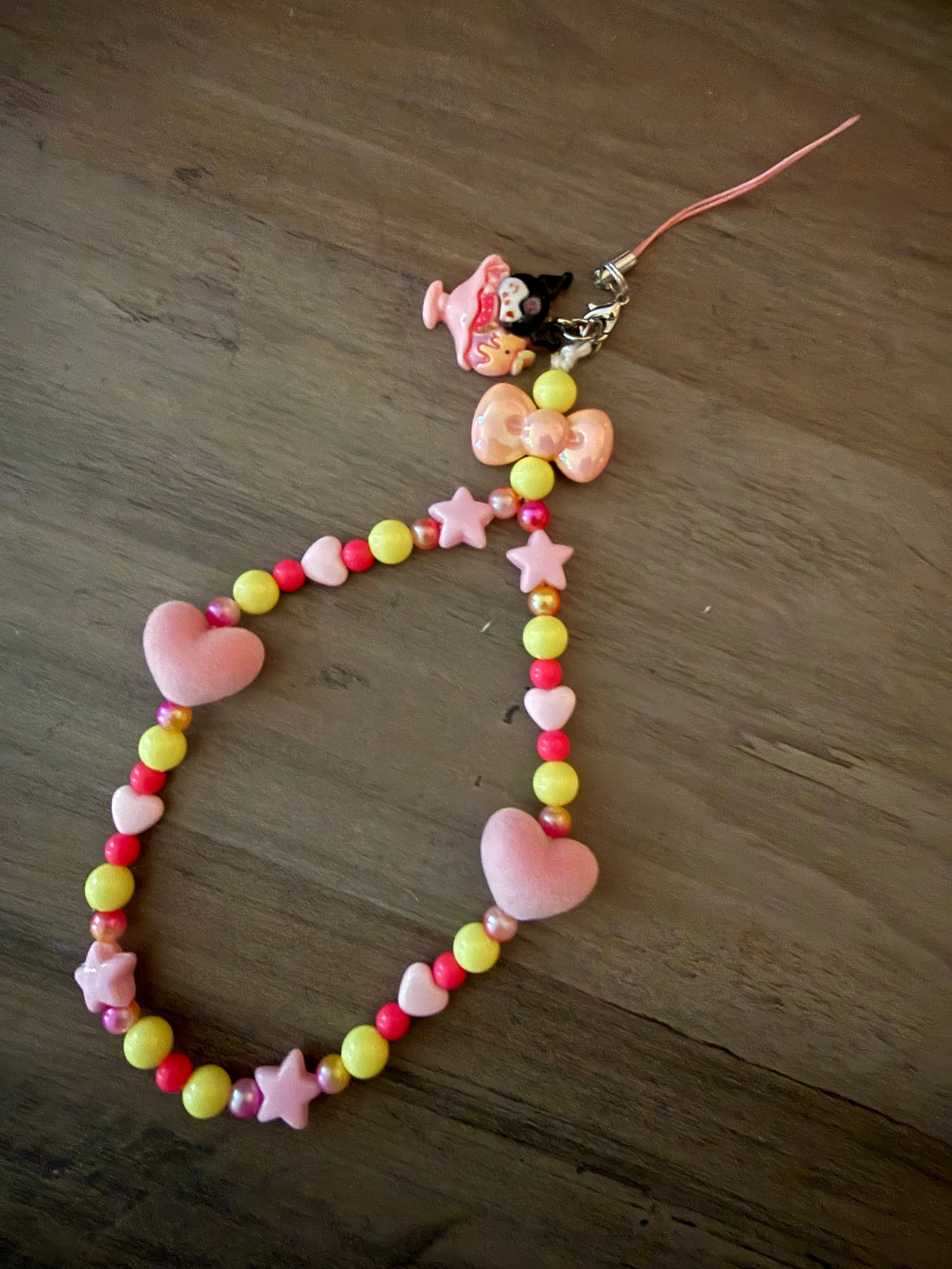 A handcrafted beaded phone wristlet featuring a mix of yellow, pink, and red beads with pink star and heart charms, accented by a peach-colored bow and a Kuromi-themed dessert charm.